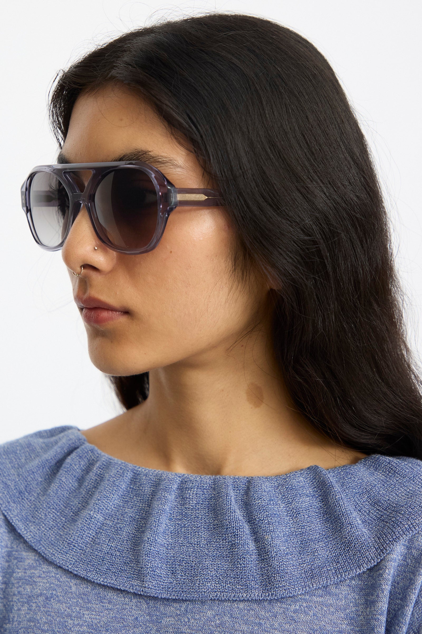 A woman with long dark hair wears Eva Masaki's Chiyo Aviator Sunglasses in Waves, featuring Italian acetate, and a blue knit top as she faces slightly left against a plain backdrop.