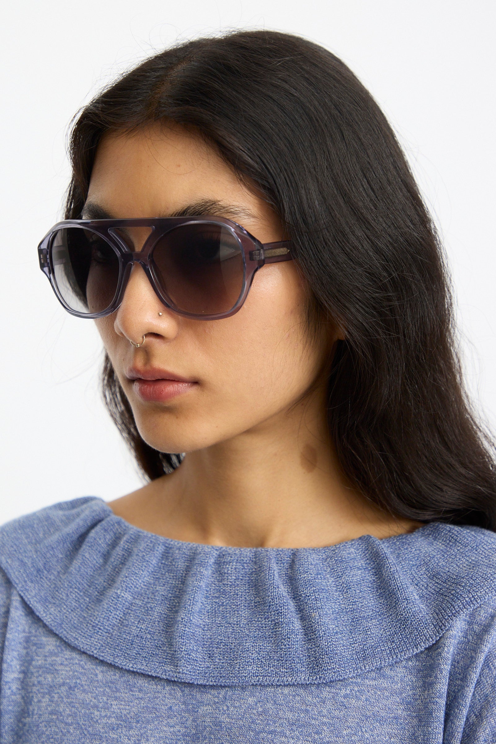 A woman with long dark hair wears Eva Masaki's Chiyo Aviator Sunglasses in Waves, crafted from Italian acetate, and a blue knit top, showcasing stylish UV protection against a plain background.