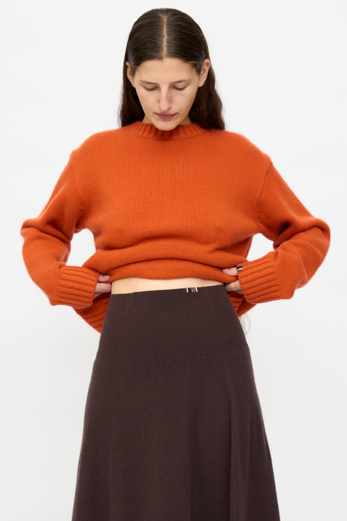 A woman models the Extreme Cashmere Ease Skirt in Dark Brown, featuring a rib-knitted waistline, paired with an orange knit sweater against a plain white background while looking down.