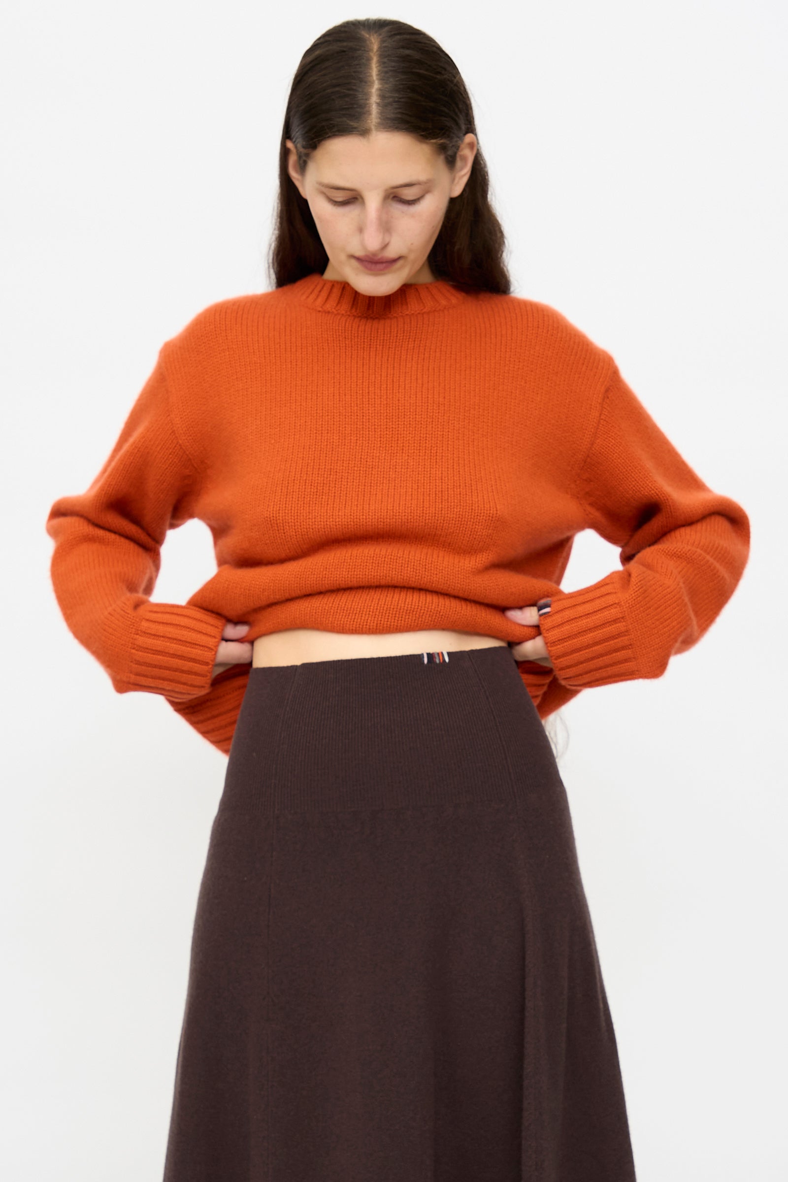 A woman models the Extreme Cashmere Ease Skirt in Dark Brown, featuring a rib-knitted waistline, paired with an orange knit sweater against a plain white background while looking down.