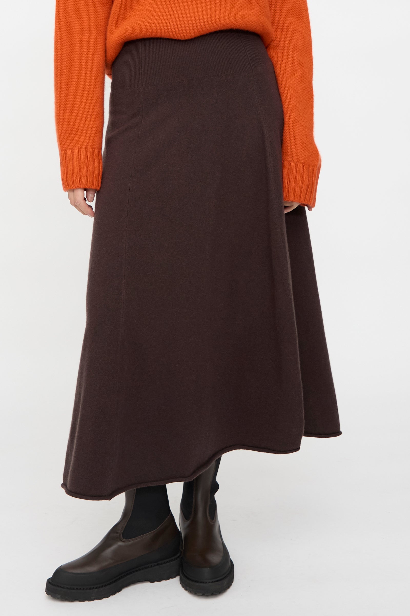 A person wearing the Ease Skirt in Dark Brown by Extreme Cashmere, paired with a burnt orange sweater, black tights, and dark brown ankle boots, stands against a plain white background.