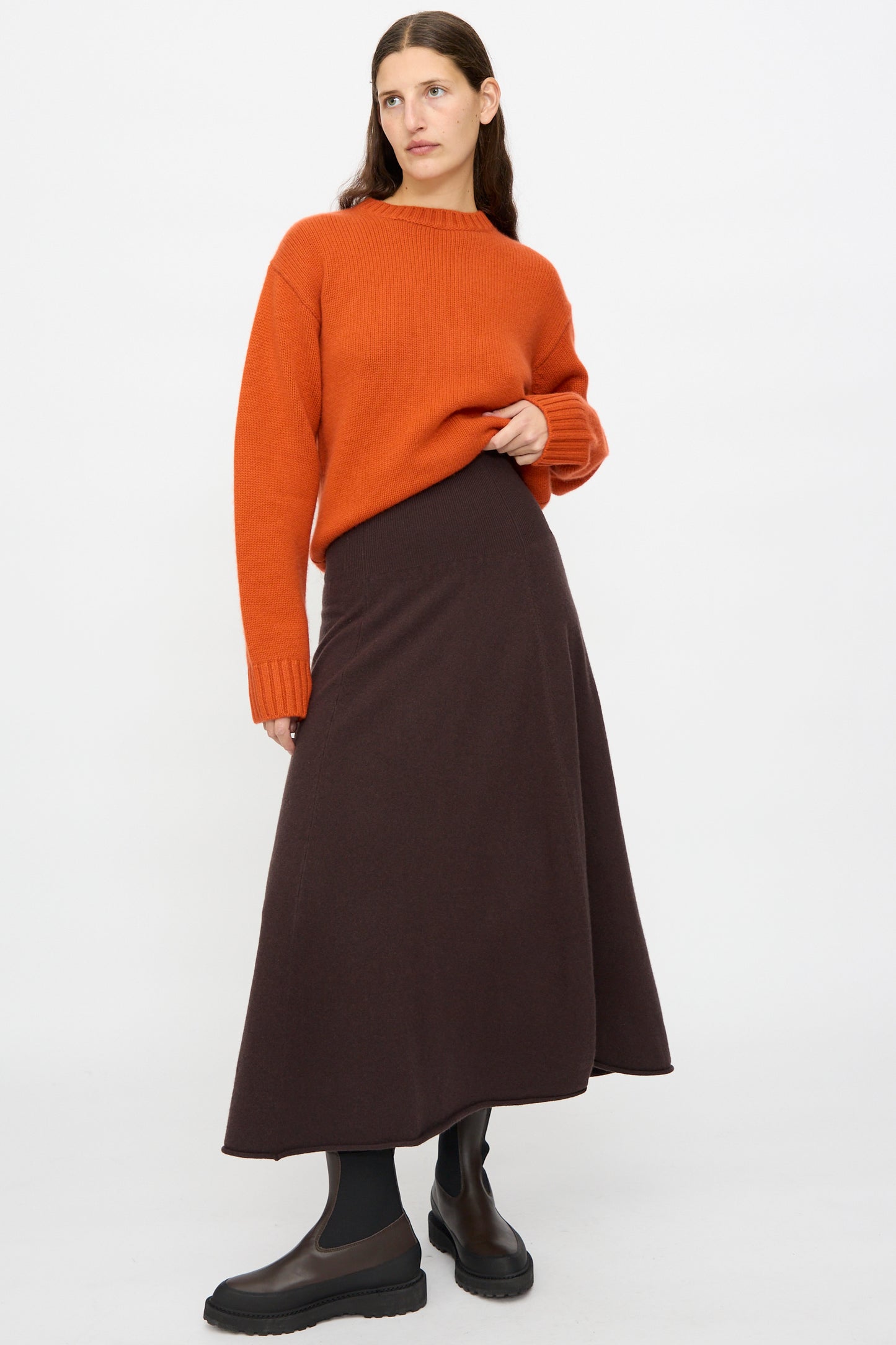 A woman wears the Extreme Cashmere Ease Skirt in Dark Brown with a rib-knitted waistline, paired with an orange sweater, black tights, and black boots, standing against a plain white background.