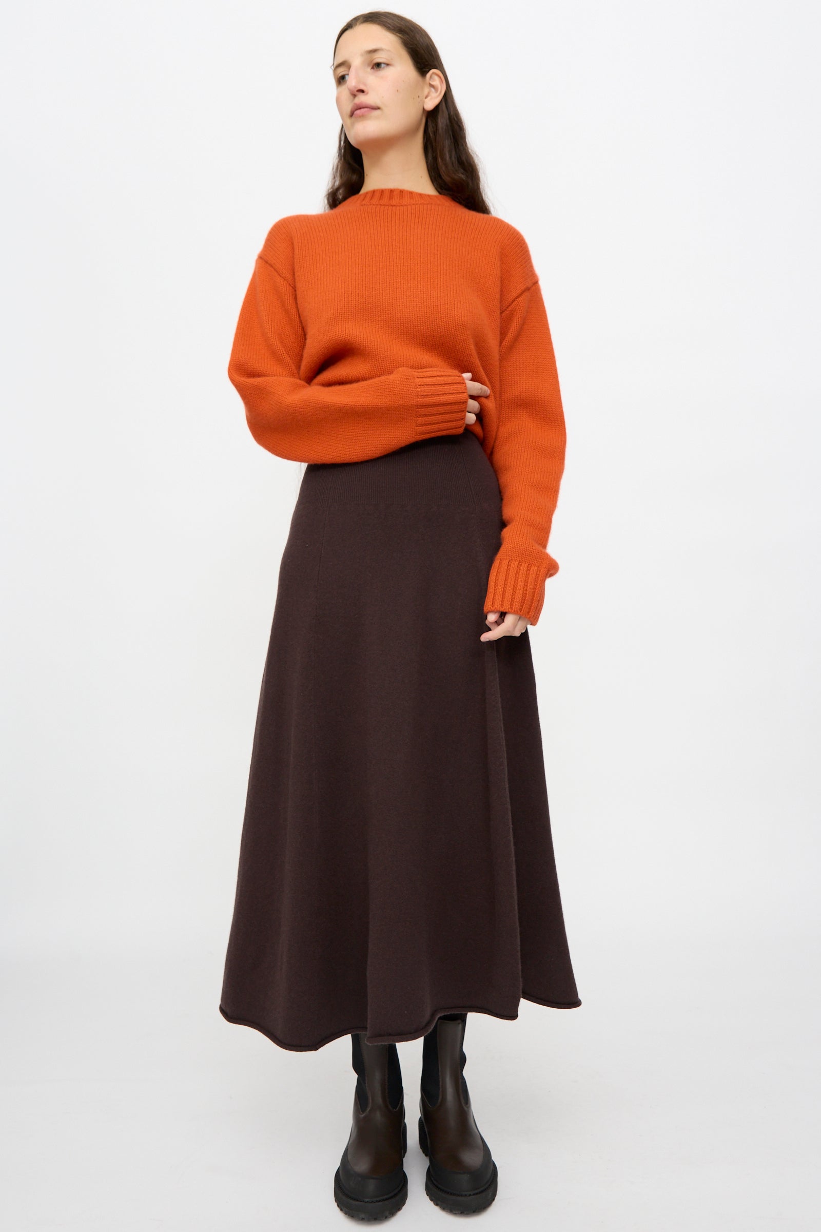 A woman stands against a white background wearing an orange sweater, black boots, and the Ease Skirt in Dark Brown by Extreme Cashmere featuring a rib-knitted waistline.