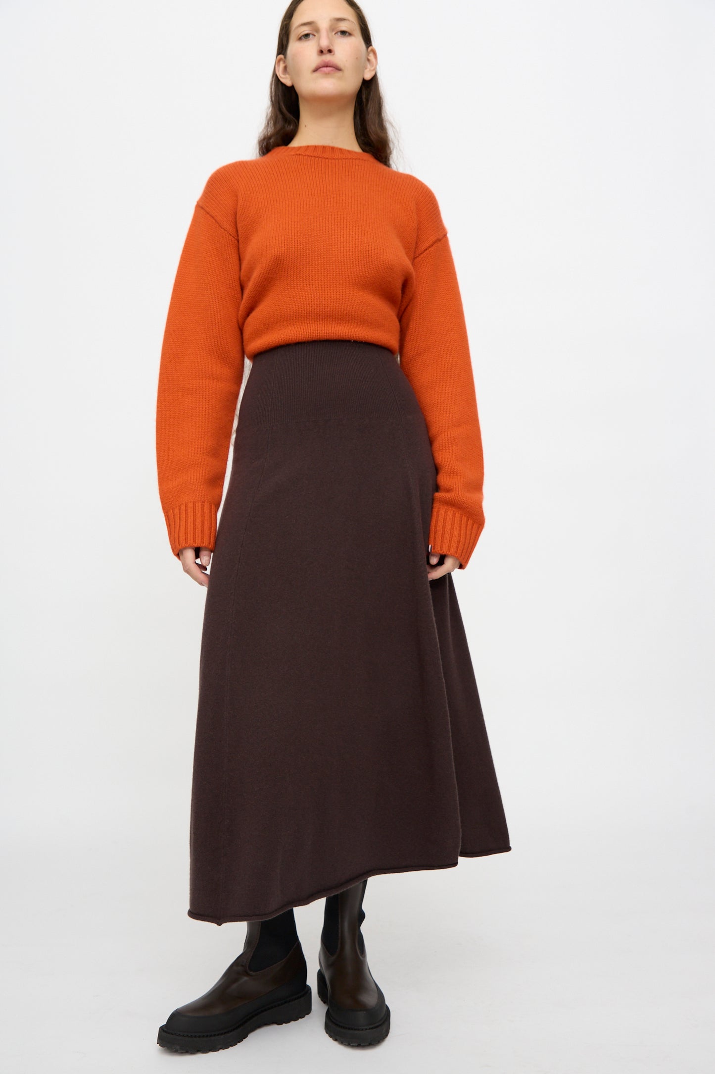 A person stands against a plain background wearing an orange sweater, black boots, and the Ease Skirt in Dark Brown by Extreme Cashmere, featuring a long silhouette and rib-knitted waistline.
