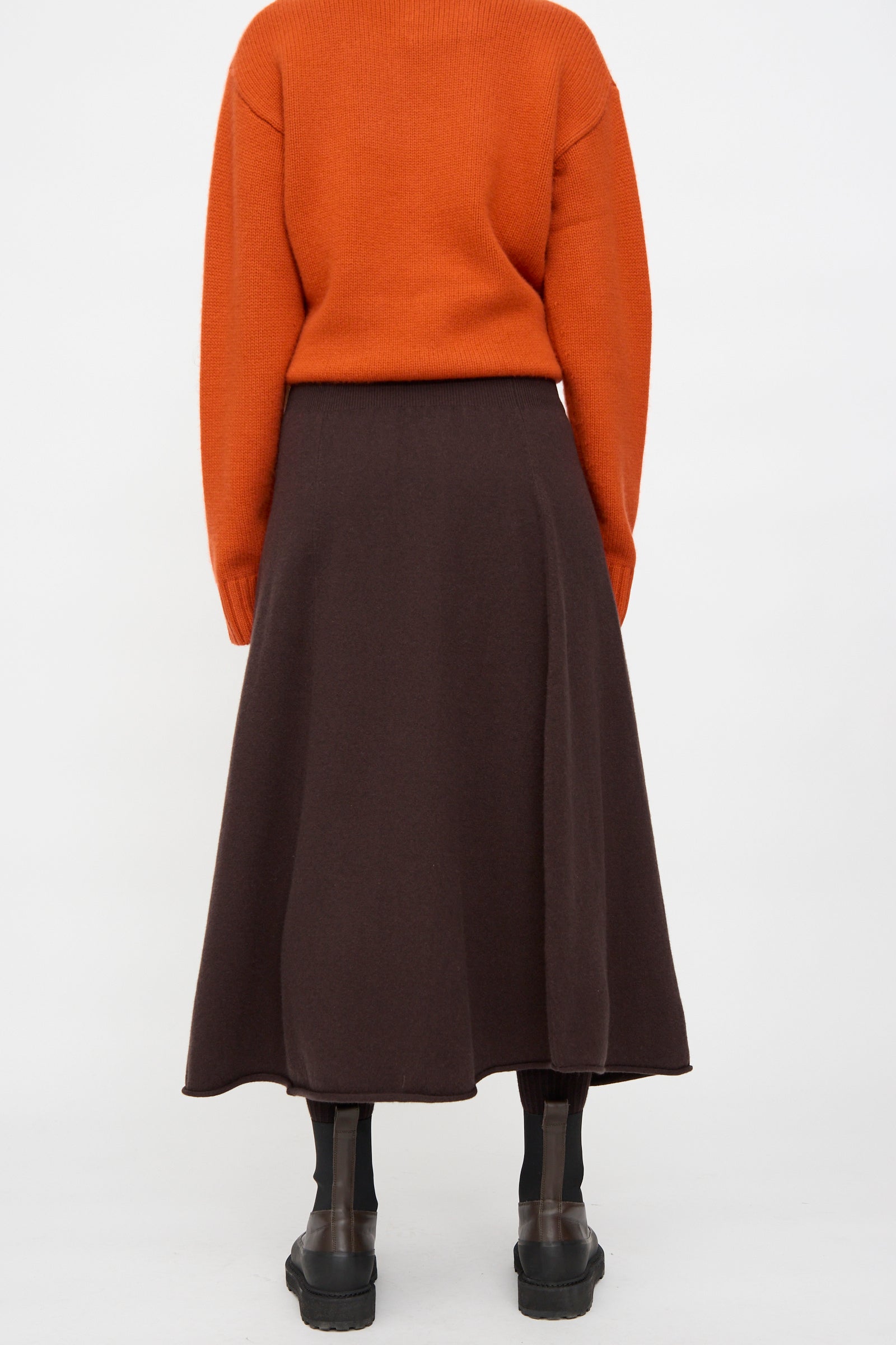 Person wearing an orange sweater, Extreme Cashmere's Ease Skirt in Dark Brown with a rib-knitted waistline, black tights, and black shoes, standing against a plain white background, photographed from the back.