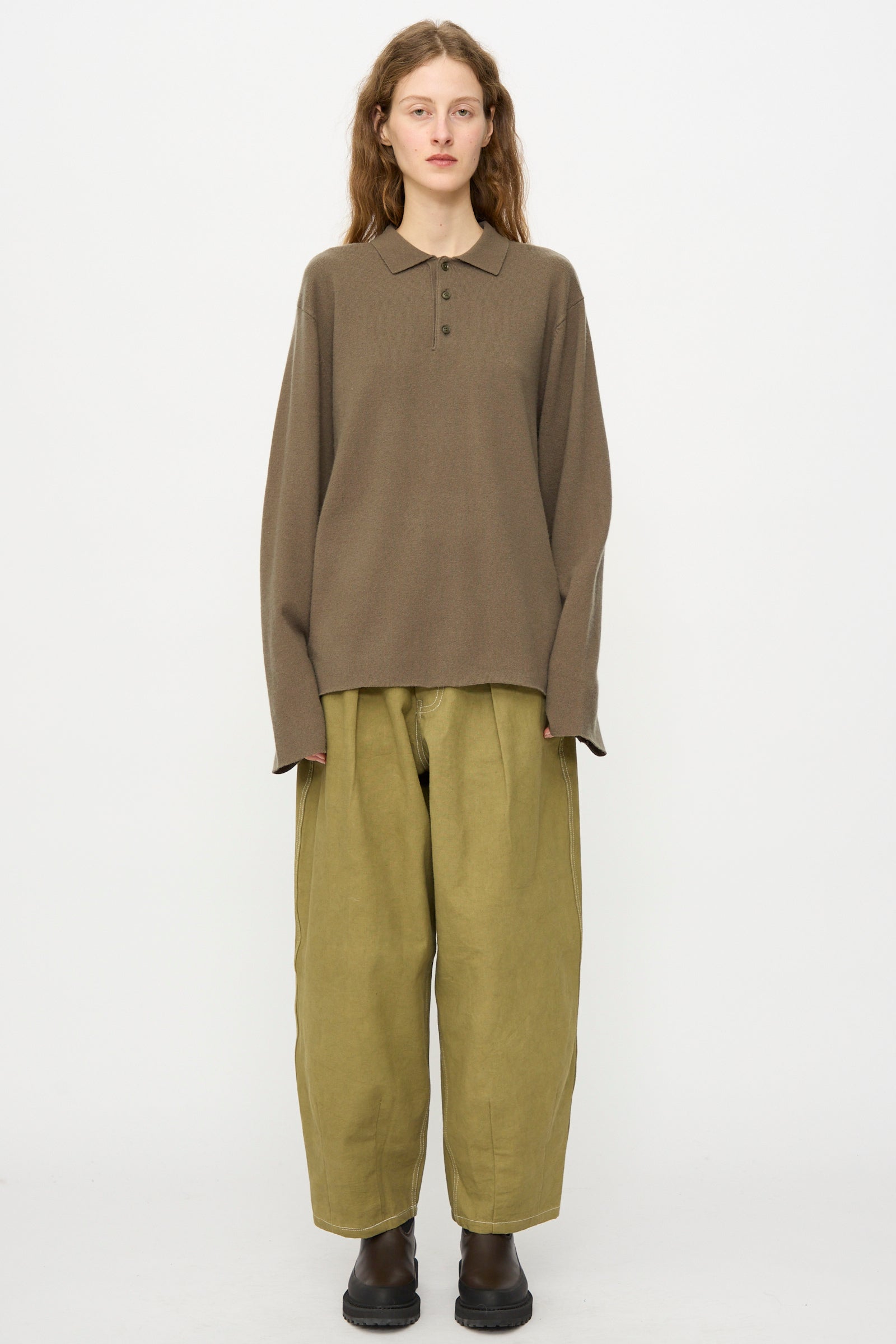 A person wears the Extreme Cashmere No. 199 Alligator Sweater in Army, paired with oversized olive green pants and dark brown boots, standing against a plain white background.