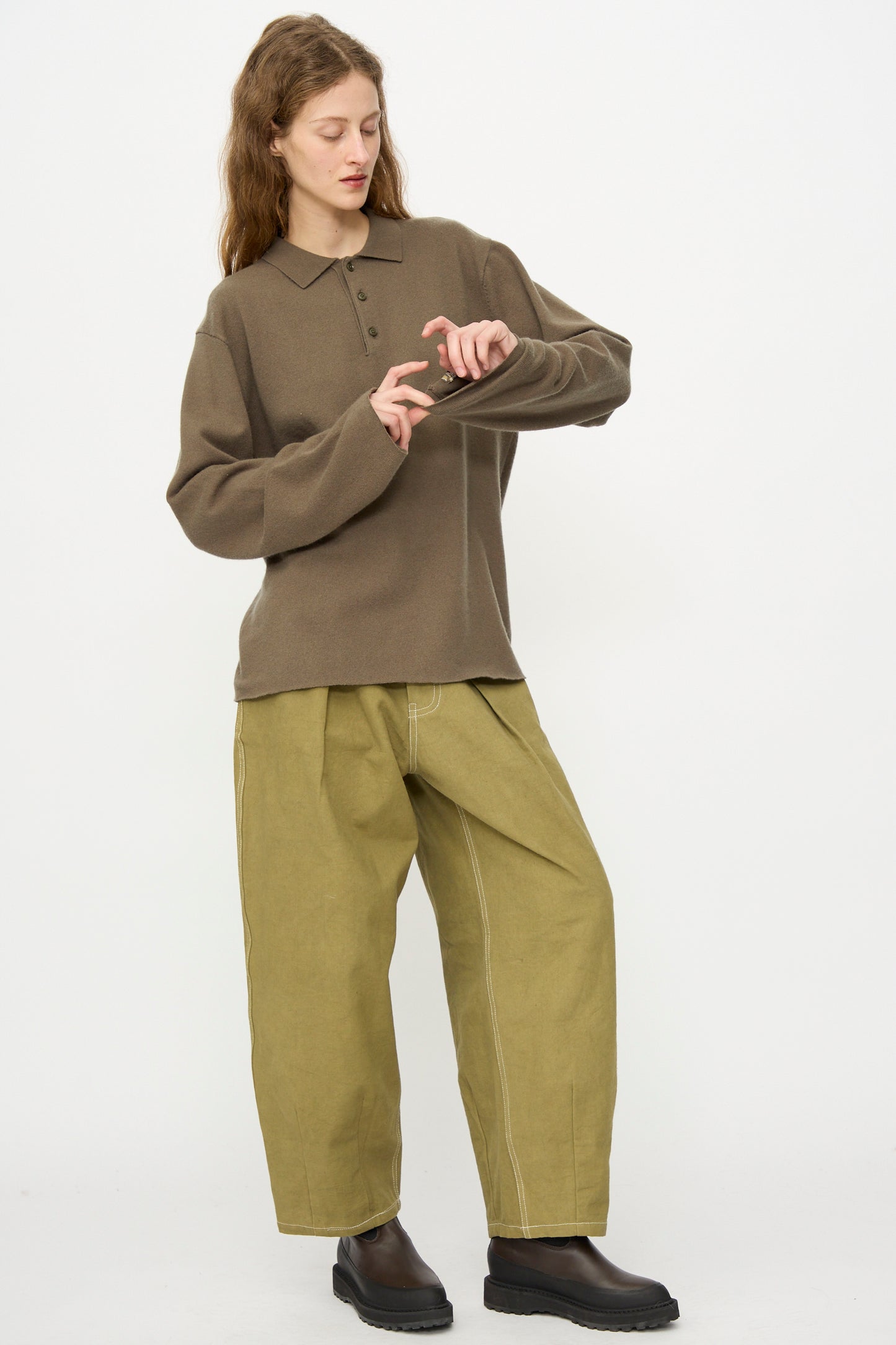 Person standing against a plain background wearing the Extreme Cashmere No. 199 Alligator Sweater in Army, paired with wide-leg light green pants and dark shoes, looking down and tapping their wrist.