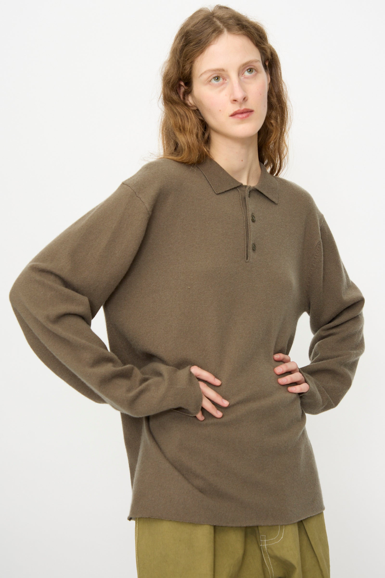 A person with long hair wears the Extreme Cashmere No. 199 Alligator Sweater in Army and olive green pants, standing against a plain white background.