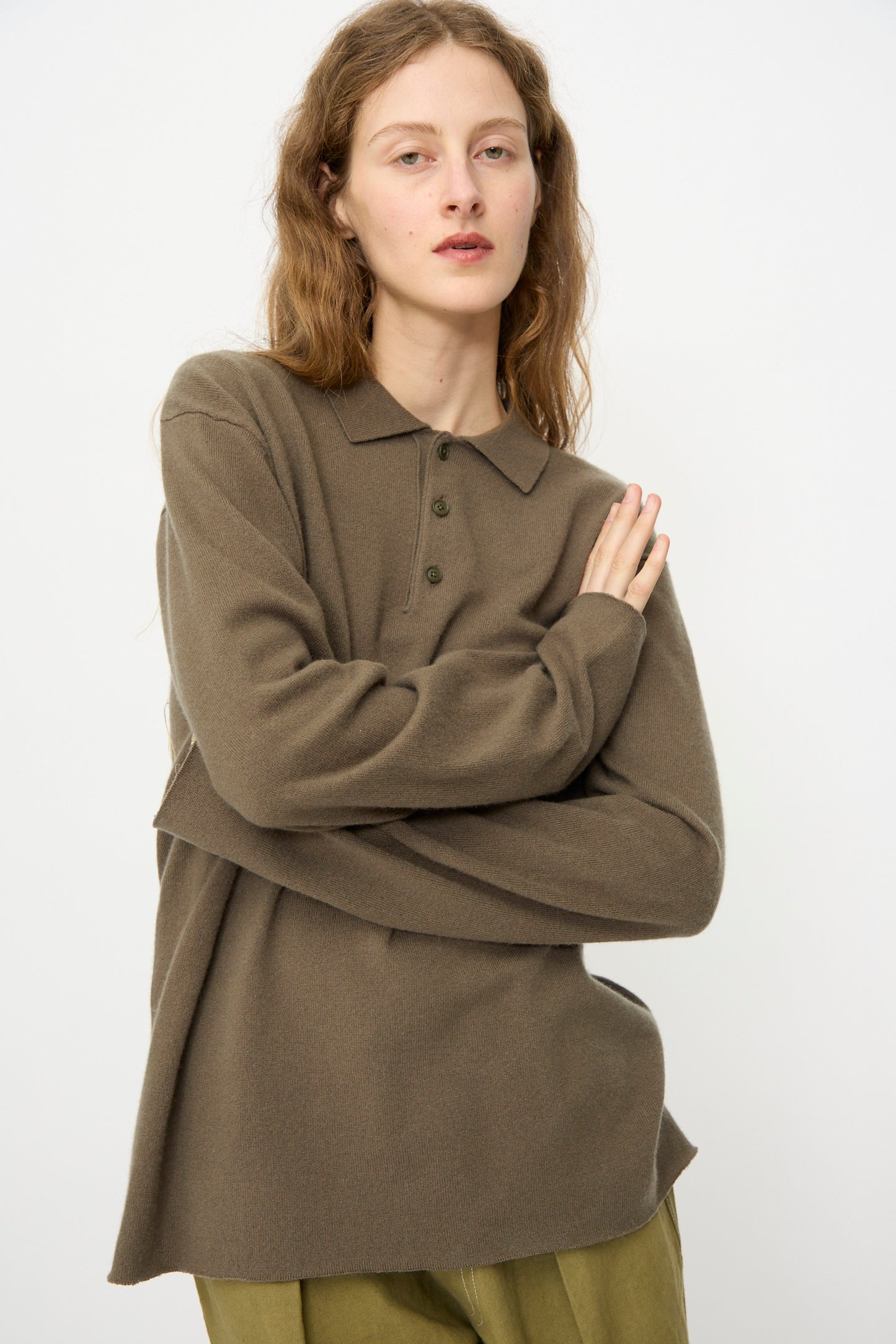 A person with long wavy hair wears the Extreme Cashmere No. 199 Alligator Sweater in Army and olive pants, standing with arms folded against a plain light background.