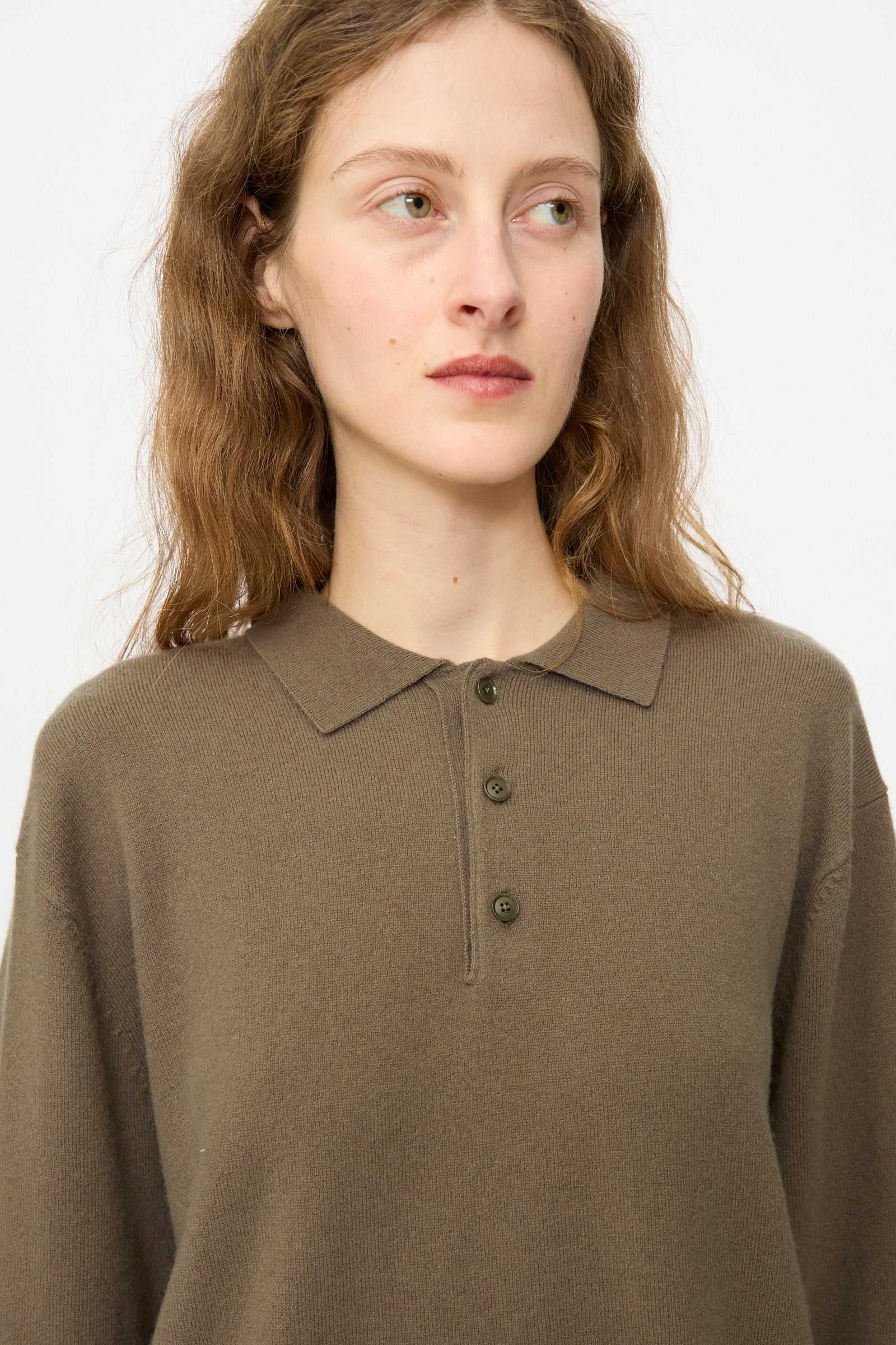 A woman with long, wavy hair wears the Extreme Cashmere No. 199 Alligator Sweater in Army, looking slightly left against a plain light background.