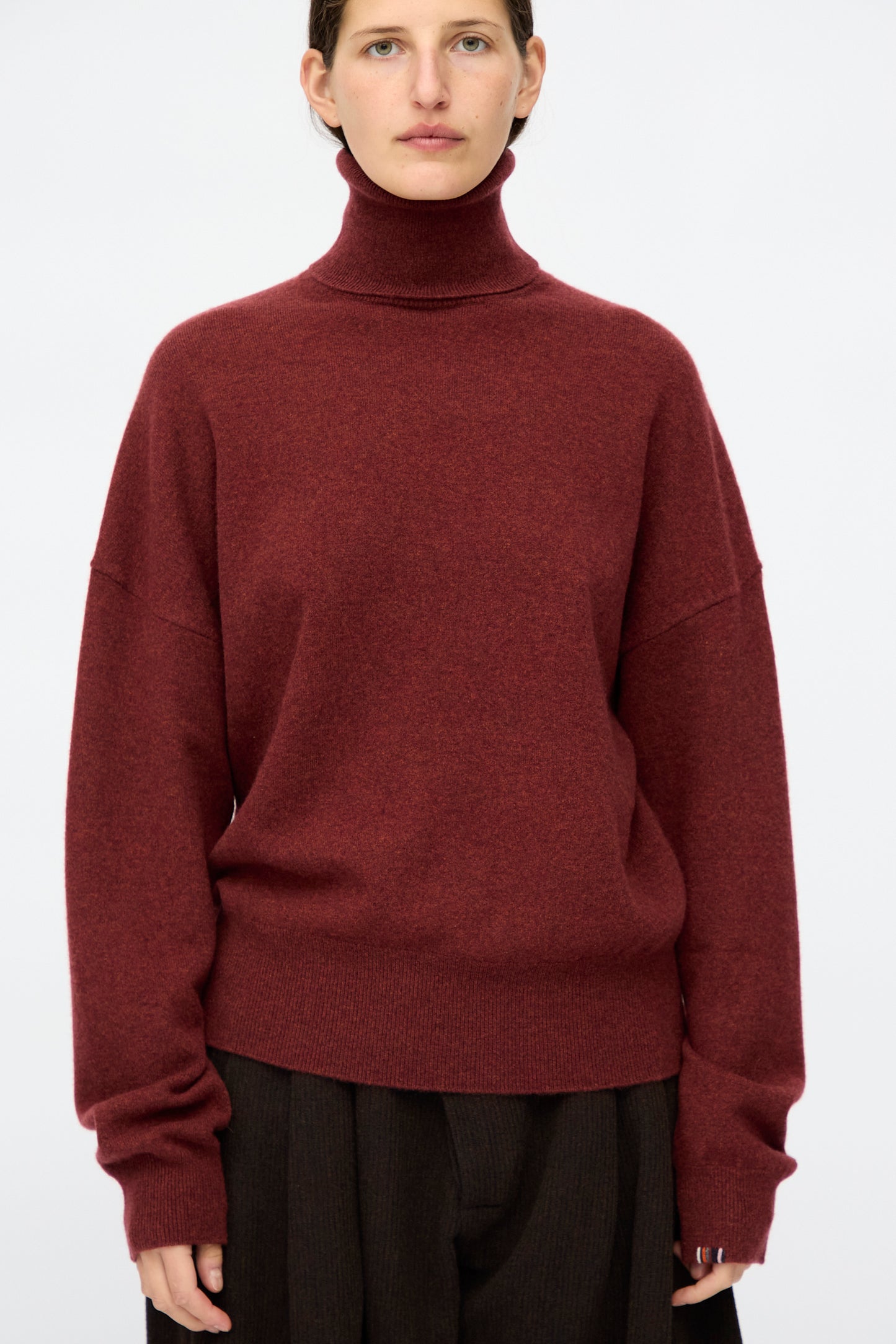 A person wearing the Extreme Cashmere No. 204 Jill Sweater in Maroon and dark trousers stands against a plain light background.