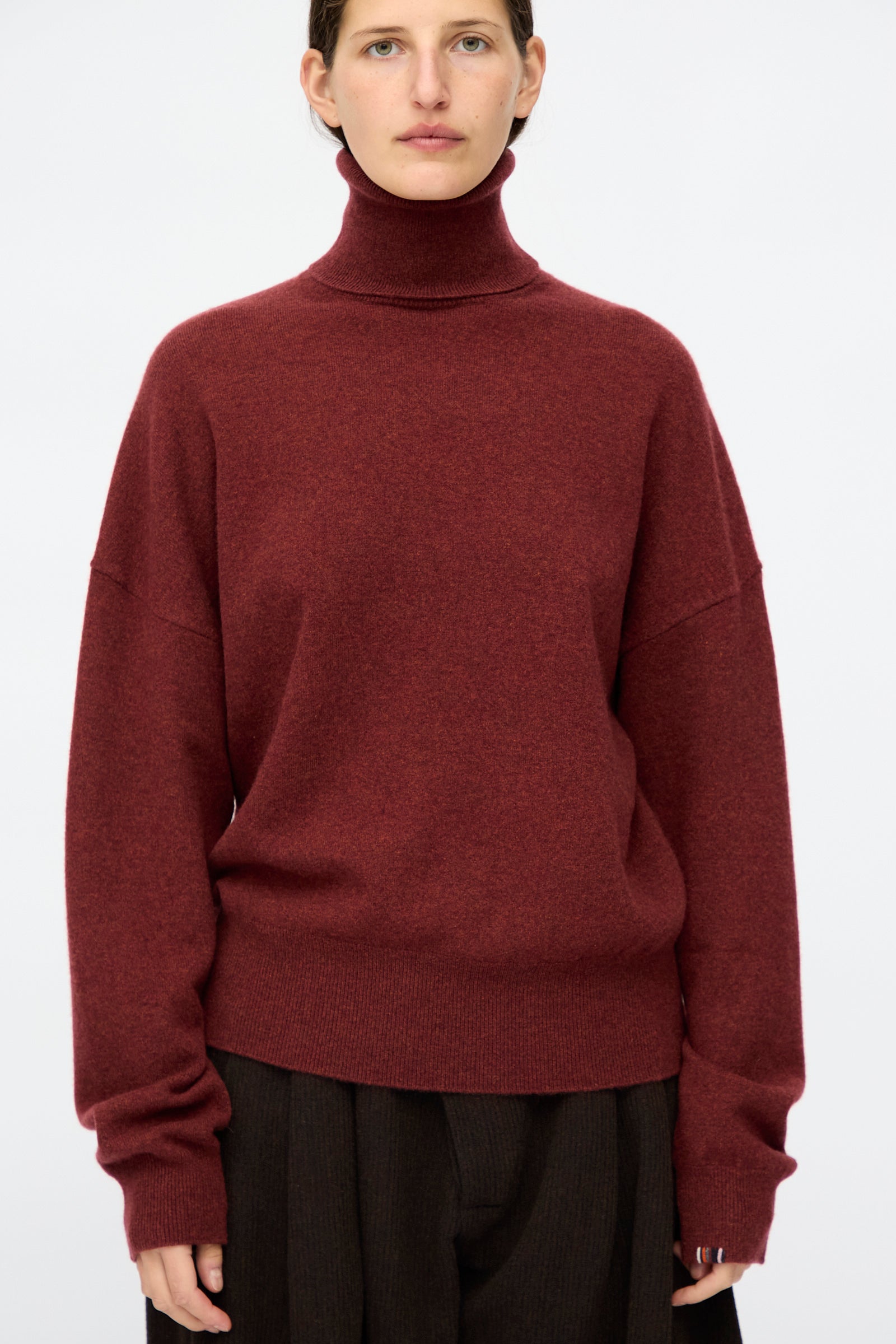 A person wearing the Extreme Cashmere No. 204 Jill Sweater in Maroon and dark trousers stands against a plain light background.