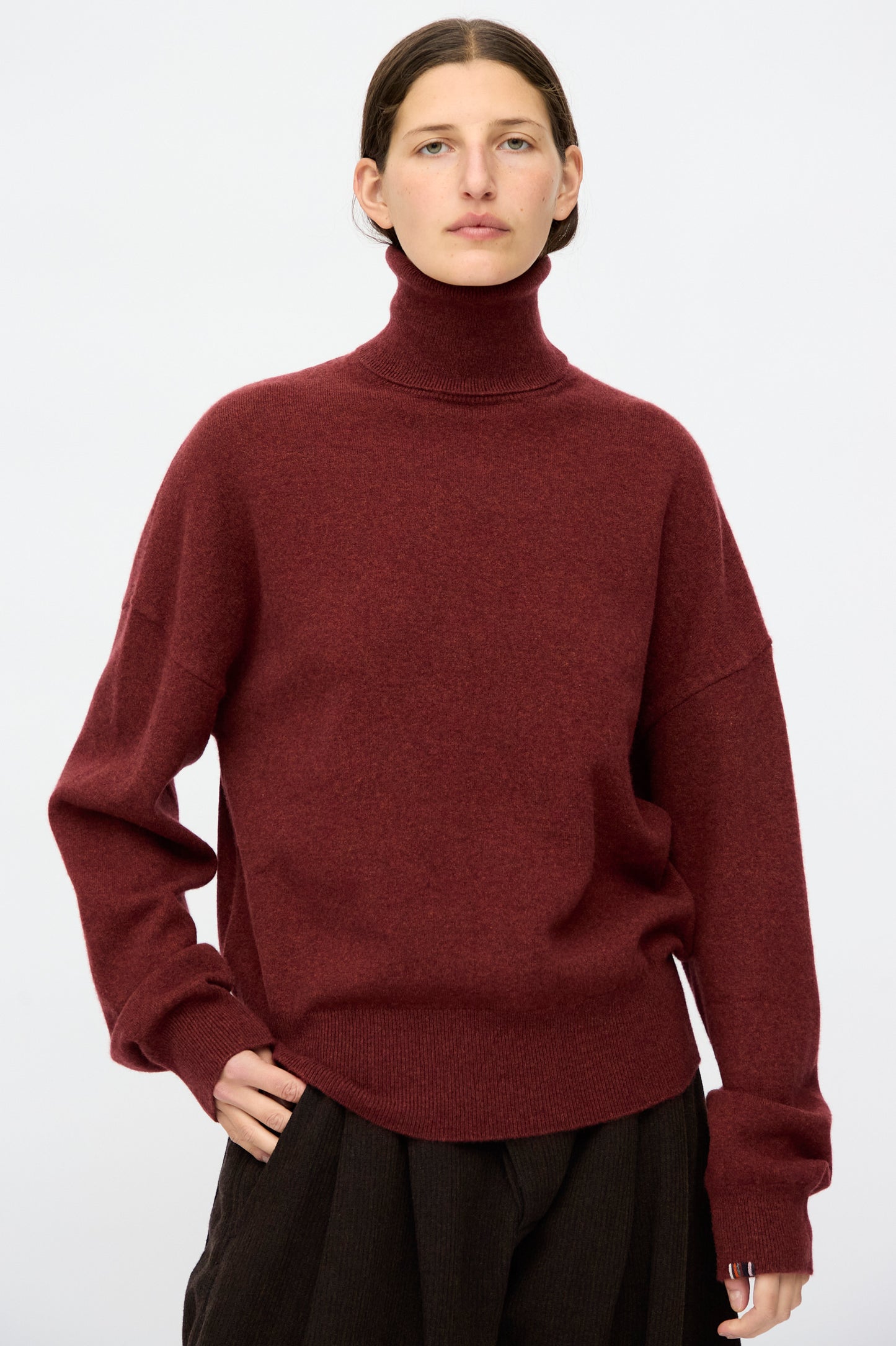 A person in the Extreme Cashmere No. 204 Jill Sweater in Maroon and dark pants stands against a white background, looking at the camera. 