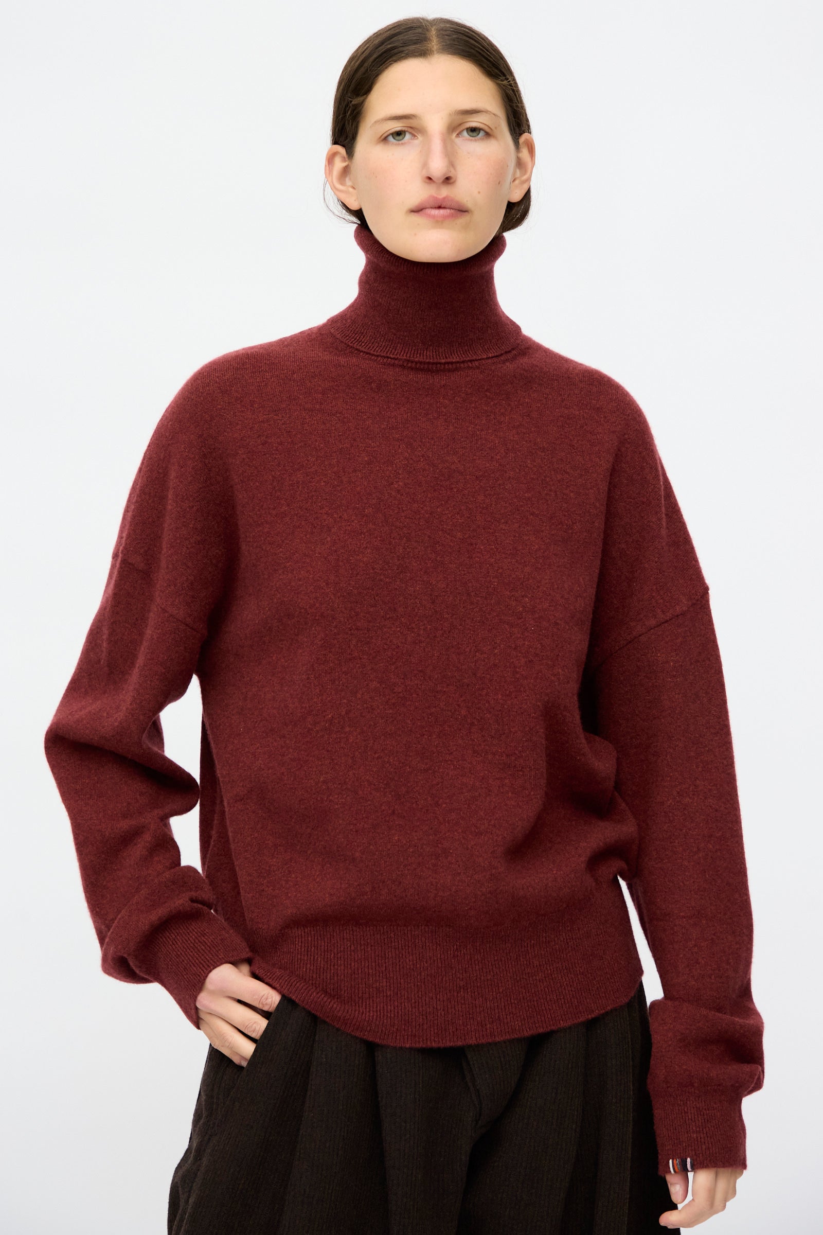 A person in the Extreme Cashmere No. 204 Jill Sweater in Maroon and dark pants stands against a white background, looking at the camera. 