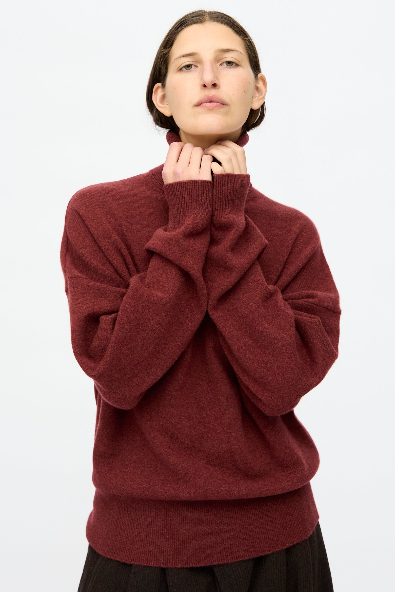 A person wearing the Extreme Cashmere No. 204 Jill Sweater in maroon, featuring long sleeves and a relaxed fit, stands against a plain light background, looking straight ahead.