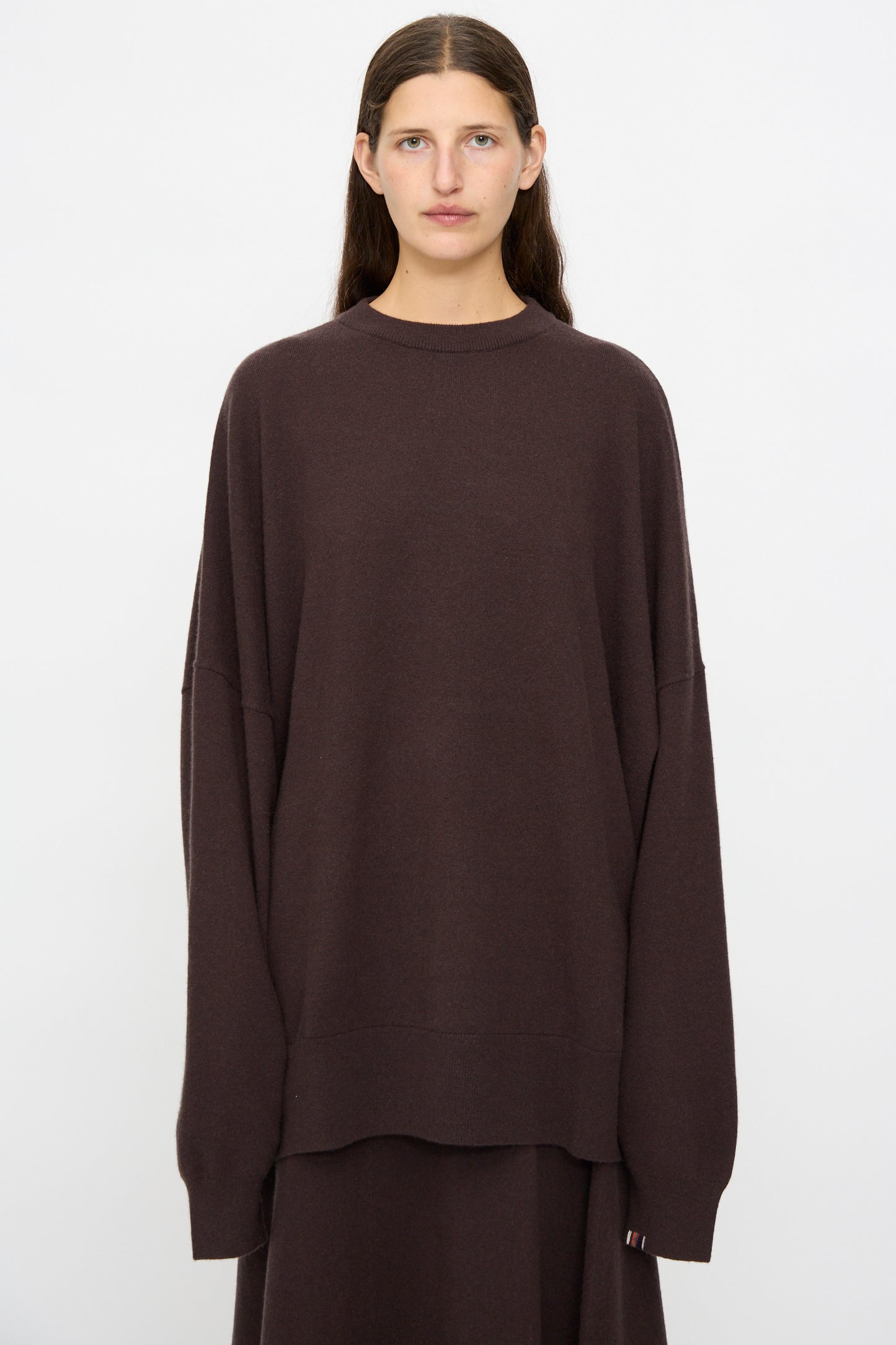 A person with long brown hair wears the Extreme Cashmere No. 246 Juna Sweater in Dark Brown with a matching skirt, standing against a plain white background.