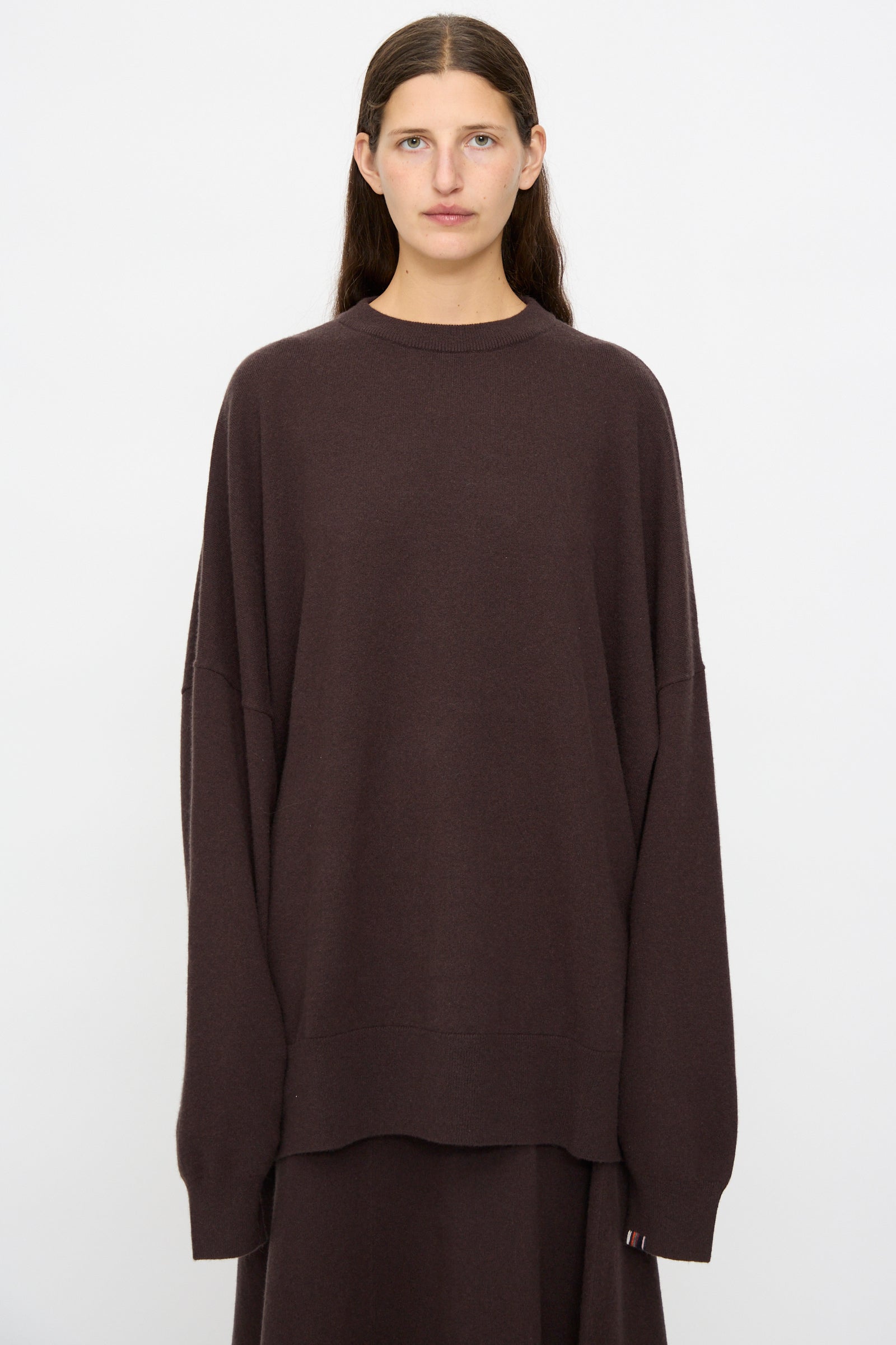 A person with long brown hair wears the Extreme Cashmere No. 246 Juna Sweater in Dark Brown with a matching skirt, standing against a plain white background.