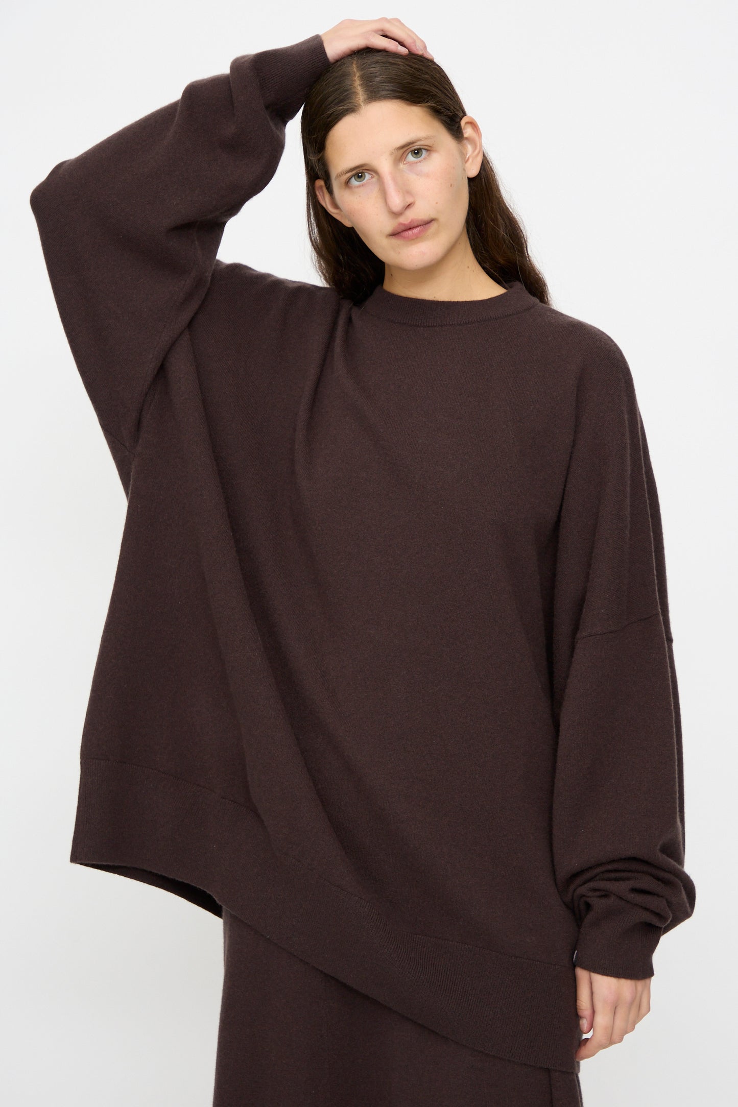 A woman wearing the Extreme Cashmere No. 246 Juna Sweater in Dark Brown, an oversized cashmere piece with long sleeves, rests one hand on her head against a plain white background.