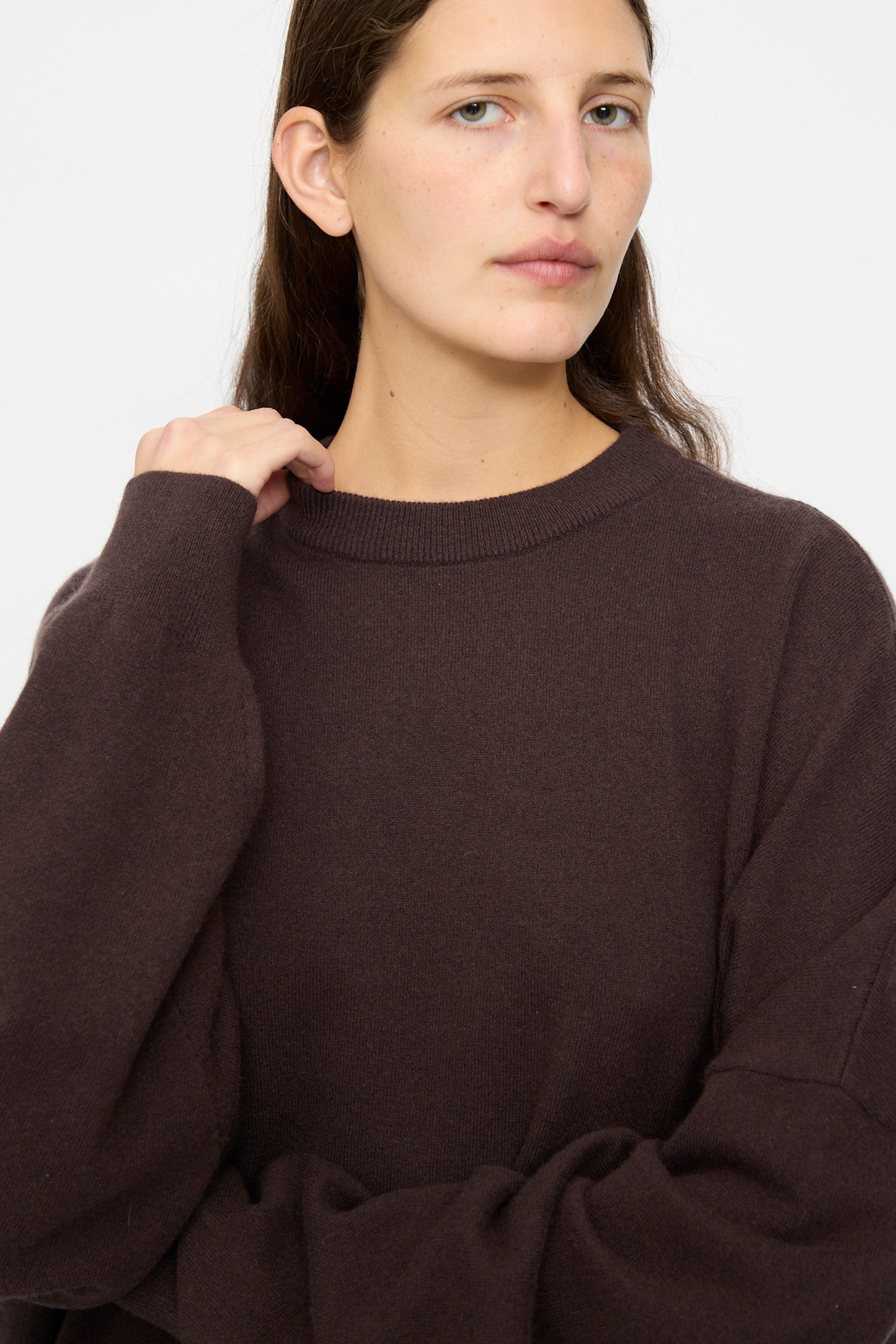 A person with long brown hair wears the No. 246 Juna Sweater in Dark Brown by Extreme Cashmere, standing against a plain background and looking slightly to the side.