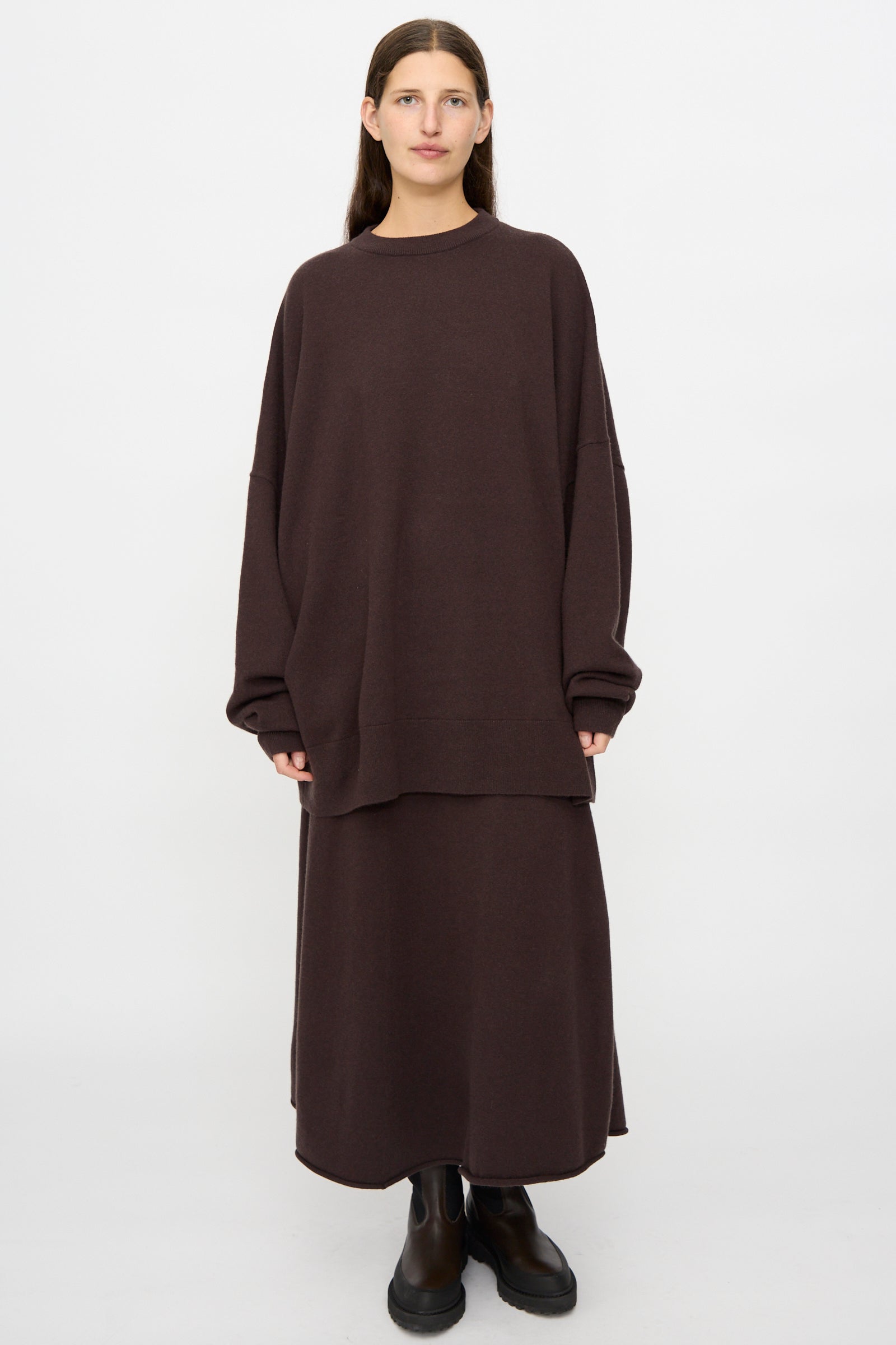 A person stands against a plain background wearing the No. 246 Juna Sweater in Dark Brown by Extreme Cashmere, paired with a matching long skirt and black boots.