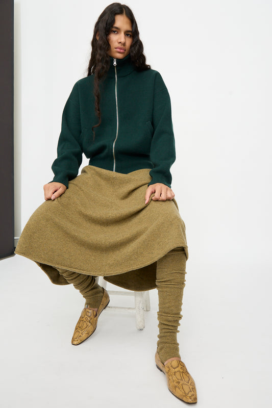 Woman wearing a dark green zip-up jacket, olive skirt, Extreme Cashmere No. 266 Ballet Socks in Hunter—rib knit cashmere leg warmers extending from knee to ankle—and snakeskin-patterned shoes, seated on a stool against a plain background.