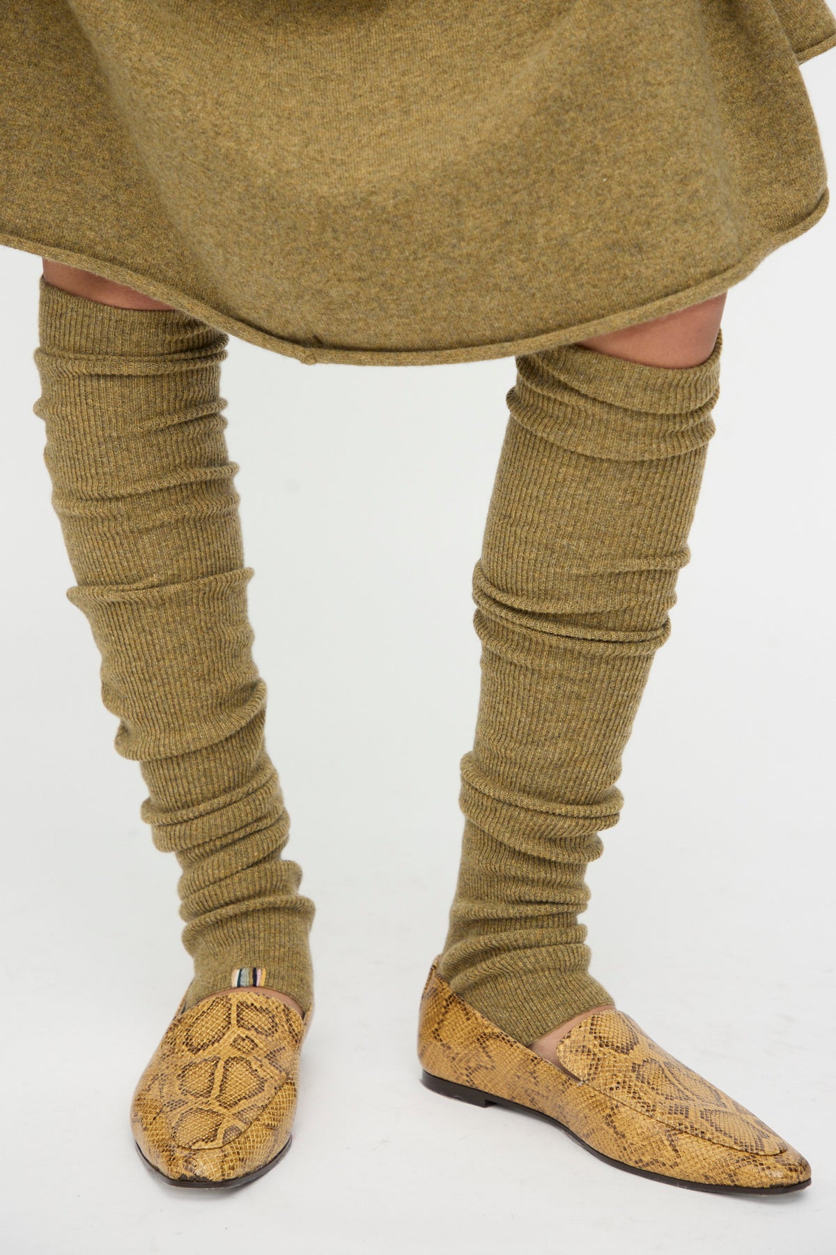 A person wearing Extreme Cashmere’s No. 266 Ballet Socks in Hunter paired with a matching skirt and tan snake-print loafers, standing on a white background.