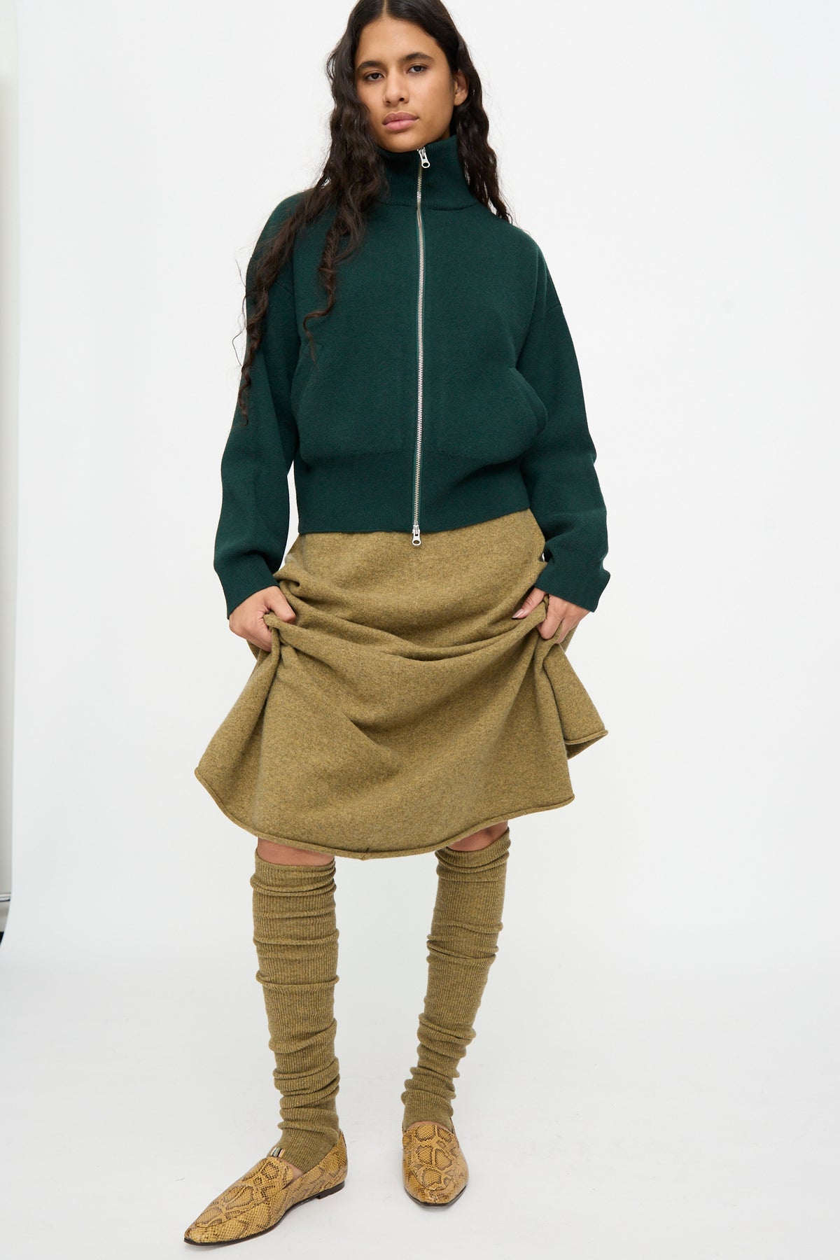 A person stands against a plain white background wearing the Extreme Cashmere No. 266 Ballet Socks in Hunter, featuring a rib knit texture that extends from knee to ankle, paired with a dark green zip-up jacket, tan skirt, and snakeskin-patterned shoes.