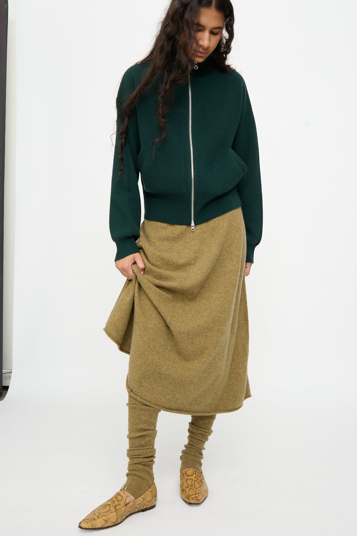 A woman stands against a white background, wearing a dark green zip-up sweater, a light olive knee to ankle length skirt with Extreme Cashmere No. 266 Ballet Socks in Hunter, and tan patterned shoes.