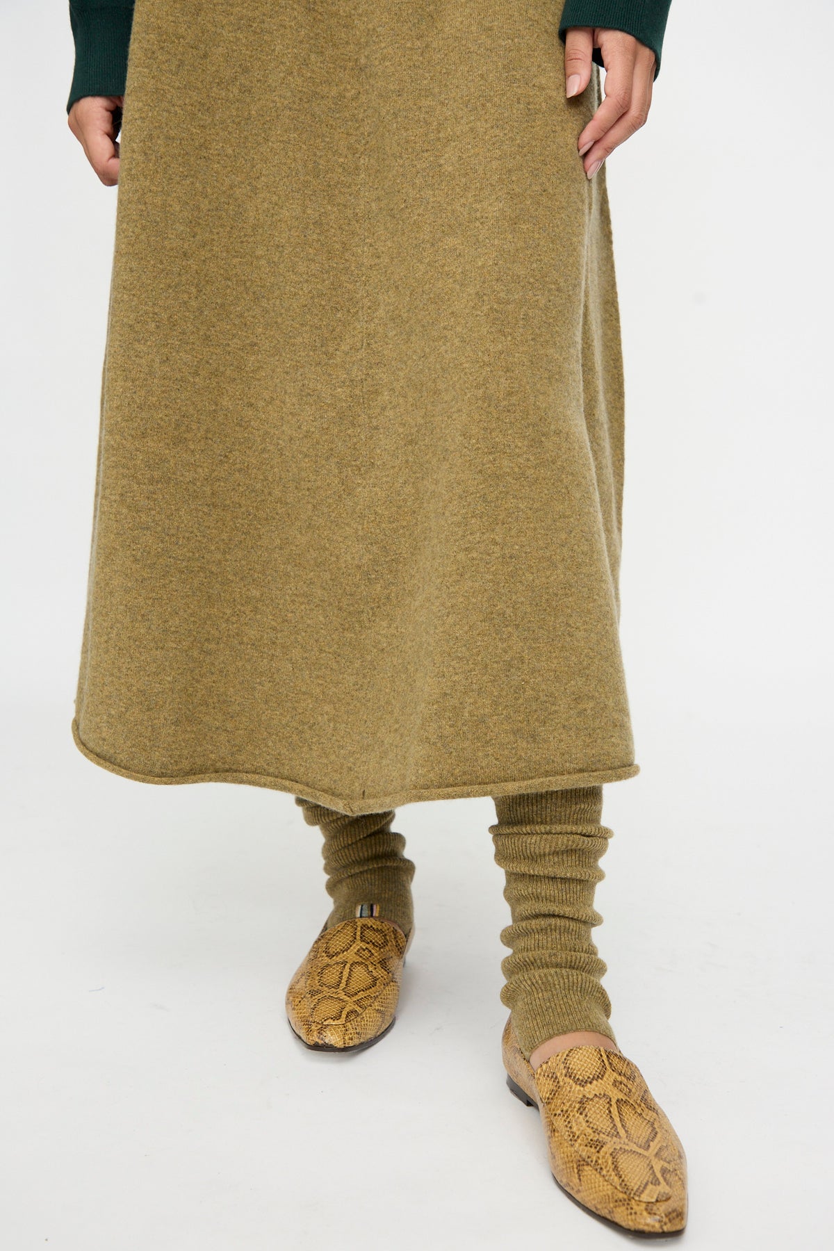 Person wearing an olive-green skirt, Extreme Cashmere’s No. 266 Ballet Socks in Hunter—rib knit cashmere leg warmers that extend from knee to ankle length—and tan snakeskin-patterned slip-on shoes, standing on a white background.