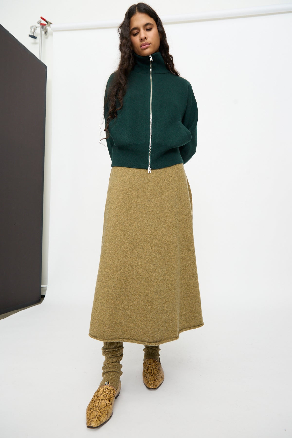 A woman stands in front of a plain studio backdrop wearing a dark green zip-up top, an olive green midi skirt, yellow snakeskin shoes, and No. 266 Ballet Socks in Hunter by Extreme Cashmere — rib knit cashmere leg warmers stretching from knee to ankle.