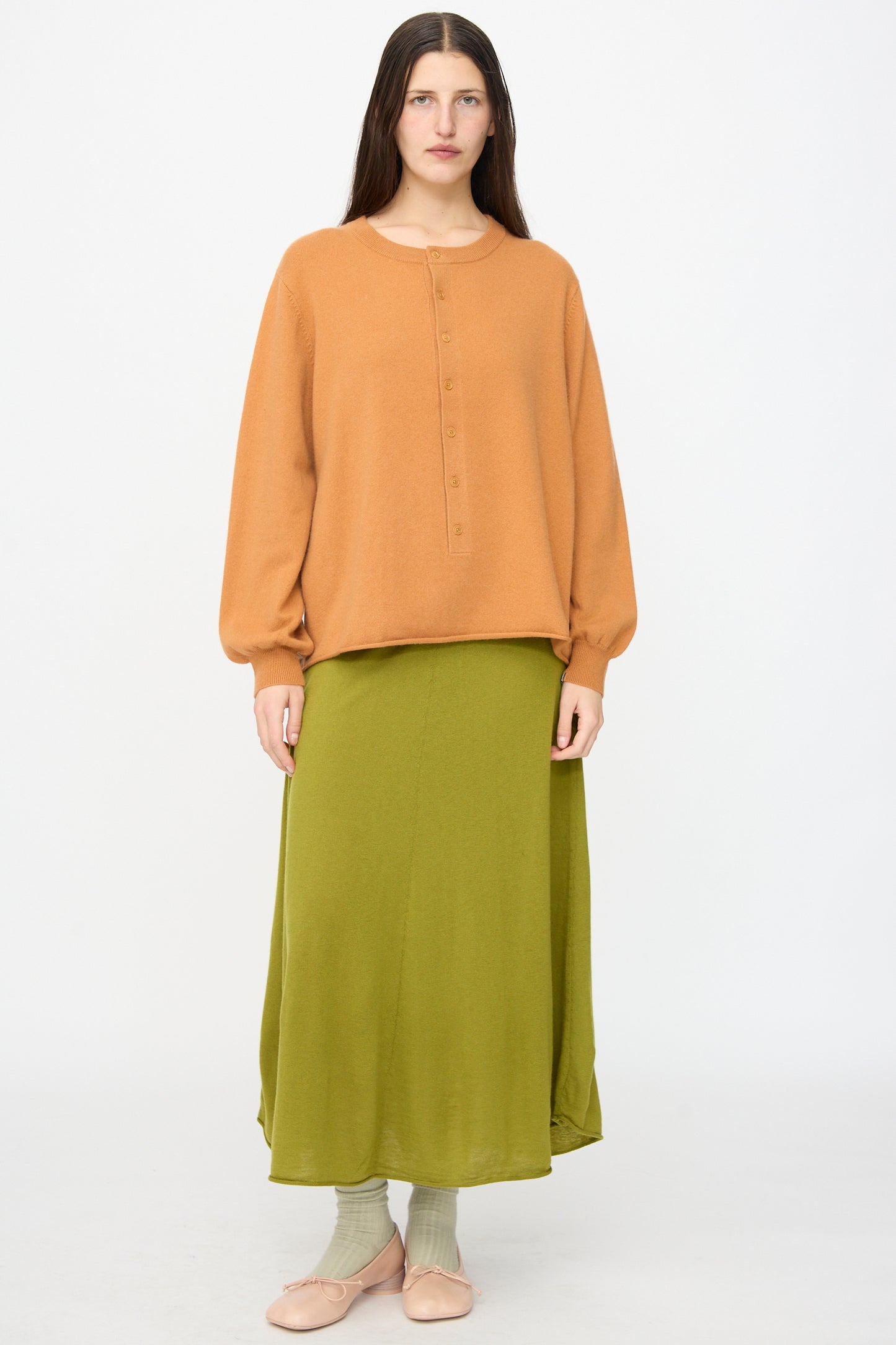 A woman stands facing forward wearing Extreme Cashmere’s No. 280 Bi Sweater in Fox, paired with an olive skirt, light green socks, and pale pink shoes against a plain white background.