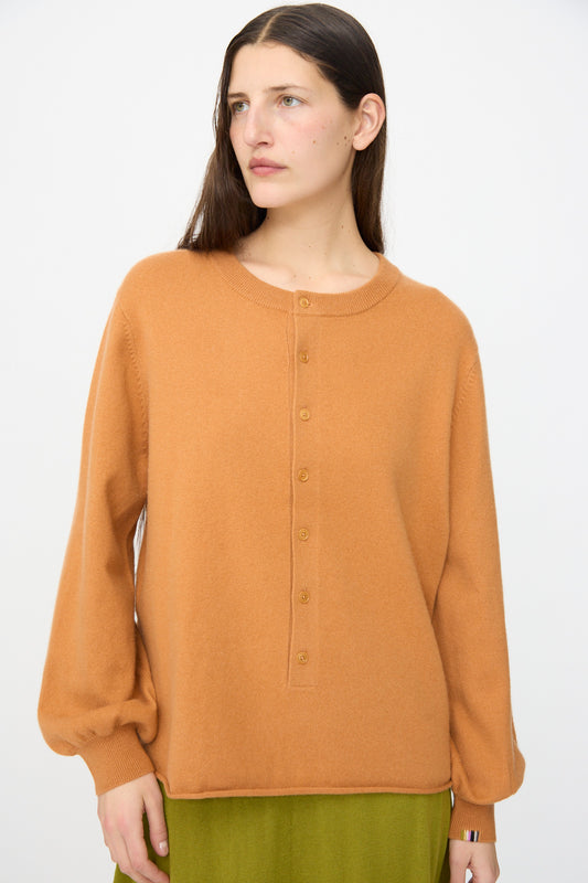 A woman with long brown hair wears the Extreme Cashmere No. 280 Bi Sweater in Fox, a camel-colored ribbed crewneck with balloon sleeves, paired with a green skirt against a plain white background.