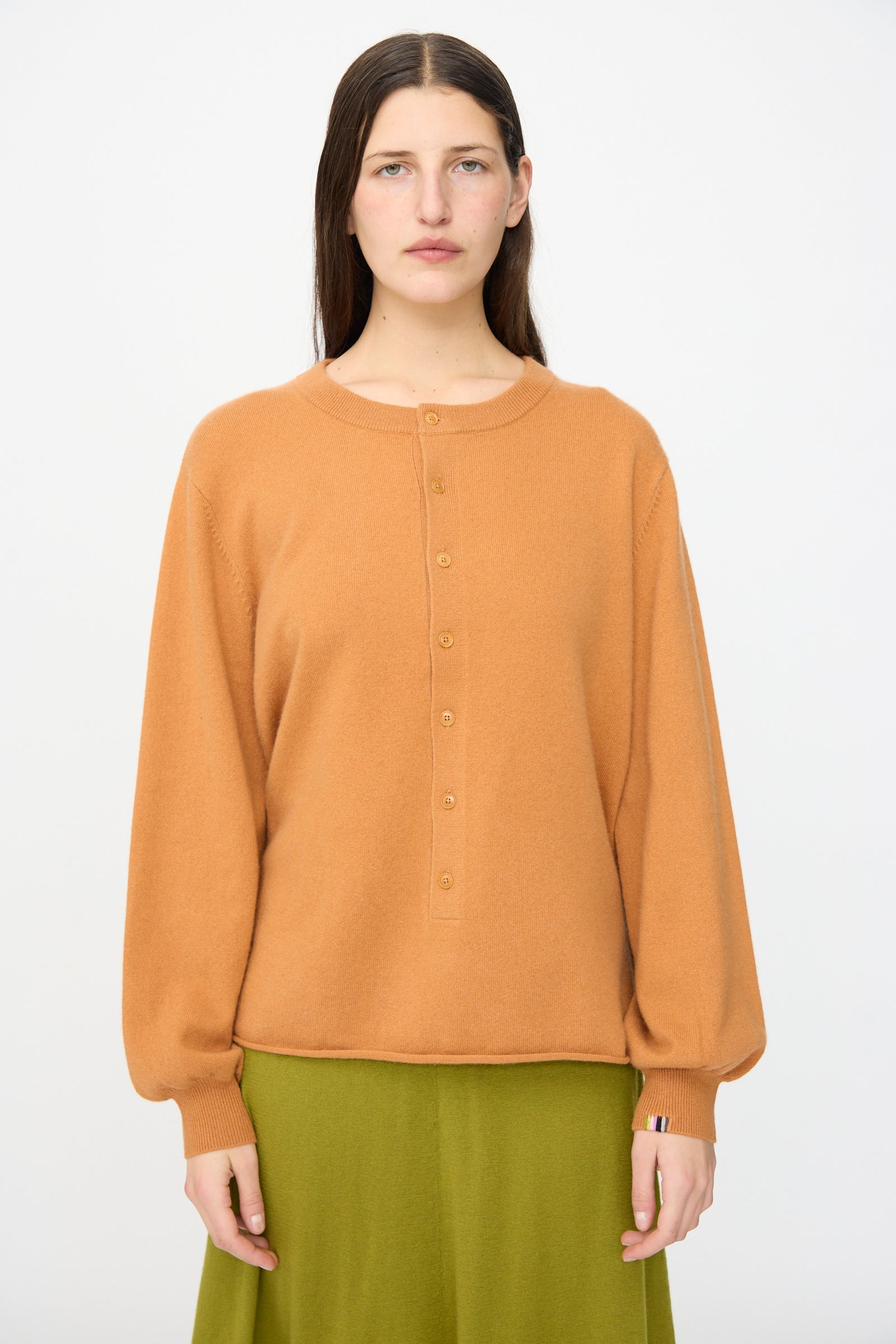 A woman with straight brown hair wears the Extreme Cashmere No. 280 Bi Sweater in Fox and an olive green skirt, standing against a plain white background.