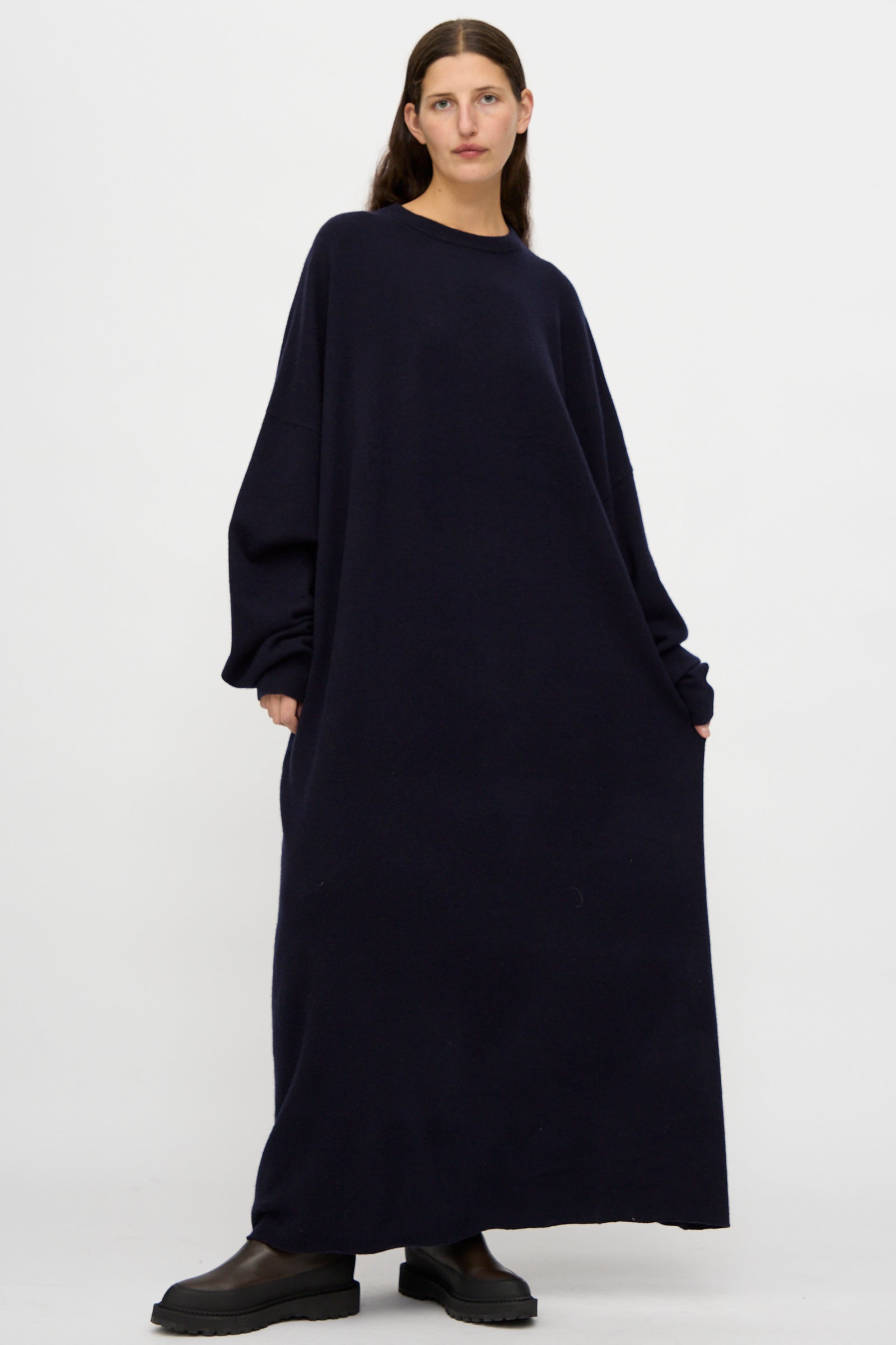 A person stands against a plain background wearing the Extreme Cashmere No. 289 May Dress in Navy, a long sleeve, loose fit cashmere maxi dress, paired with black boots.