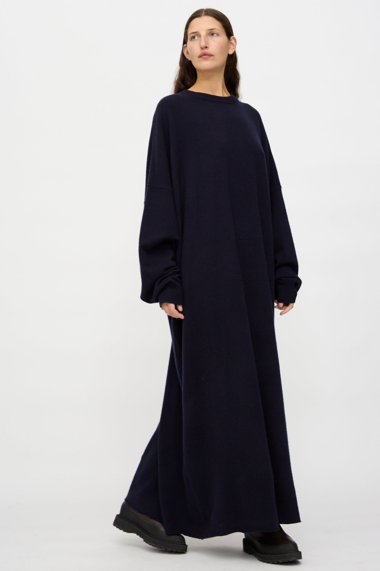 A woman stands against a plain background wearing Extreme Cashmere's No. 289 May Dress in Navy, a loose fit, long sleeve cashmere maxi, paired with black shoes.