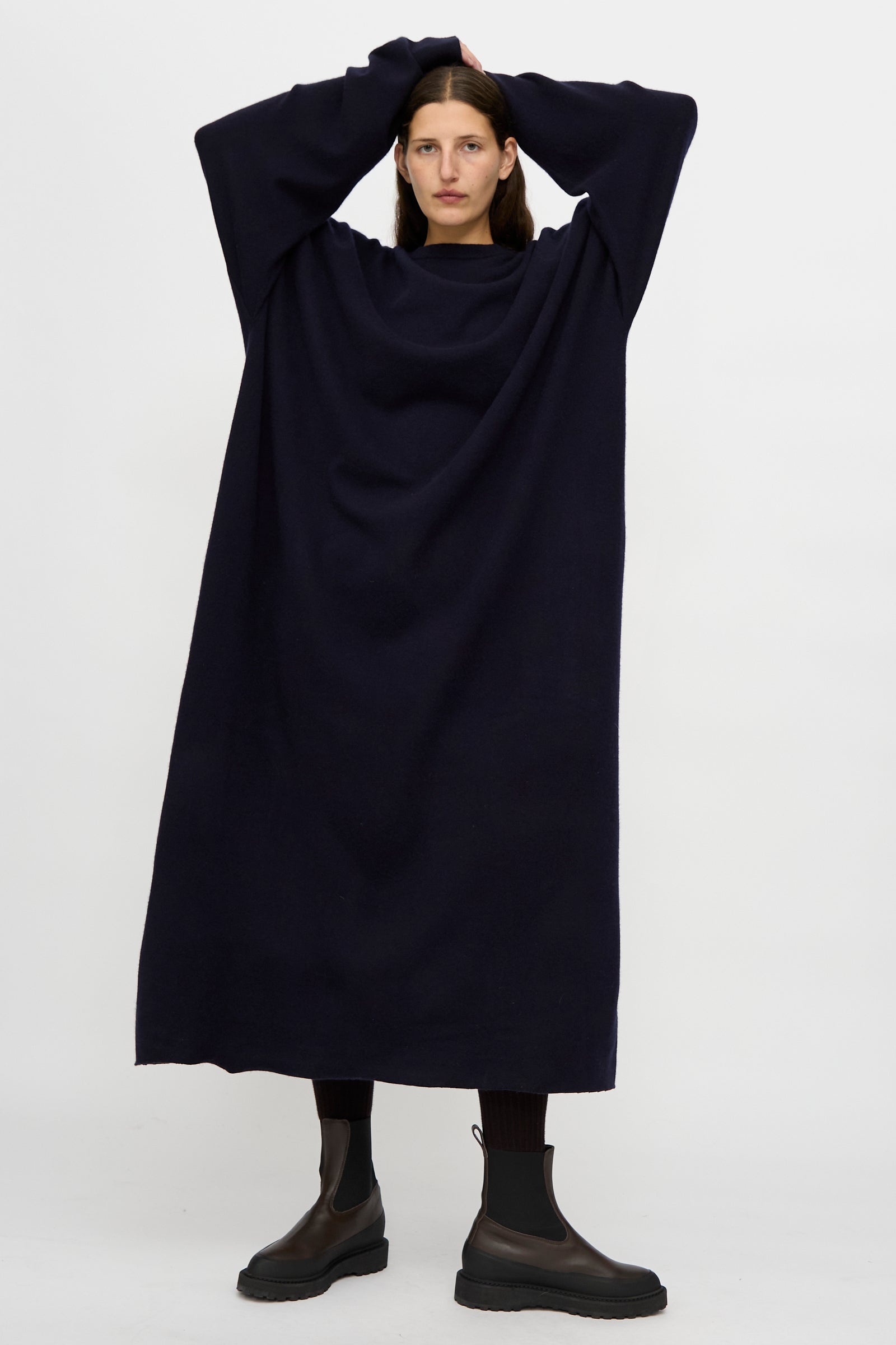 A person stands against a plain background wearing the Extreme Cashmere No. 289 May Dress in Navy, a loose fit, long sleeve dark dress, paired with black boots and resting their hands on their head.
