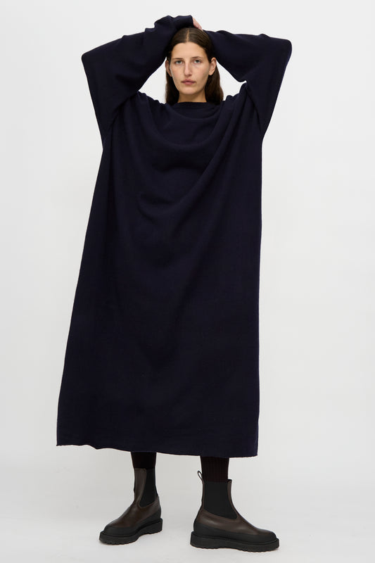 A person stands against a plain background wearing the Extreme Cashmere No. 289 May Dress in Navy, a loose fit, long sleeve dark dress, paired with black boots and resting their hands on their head.