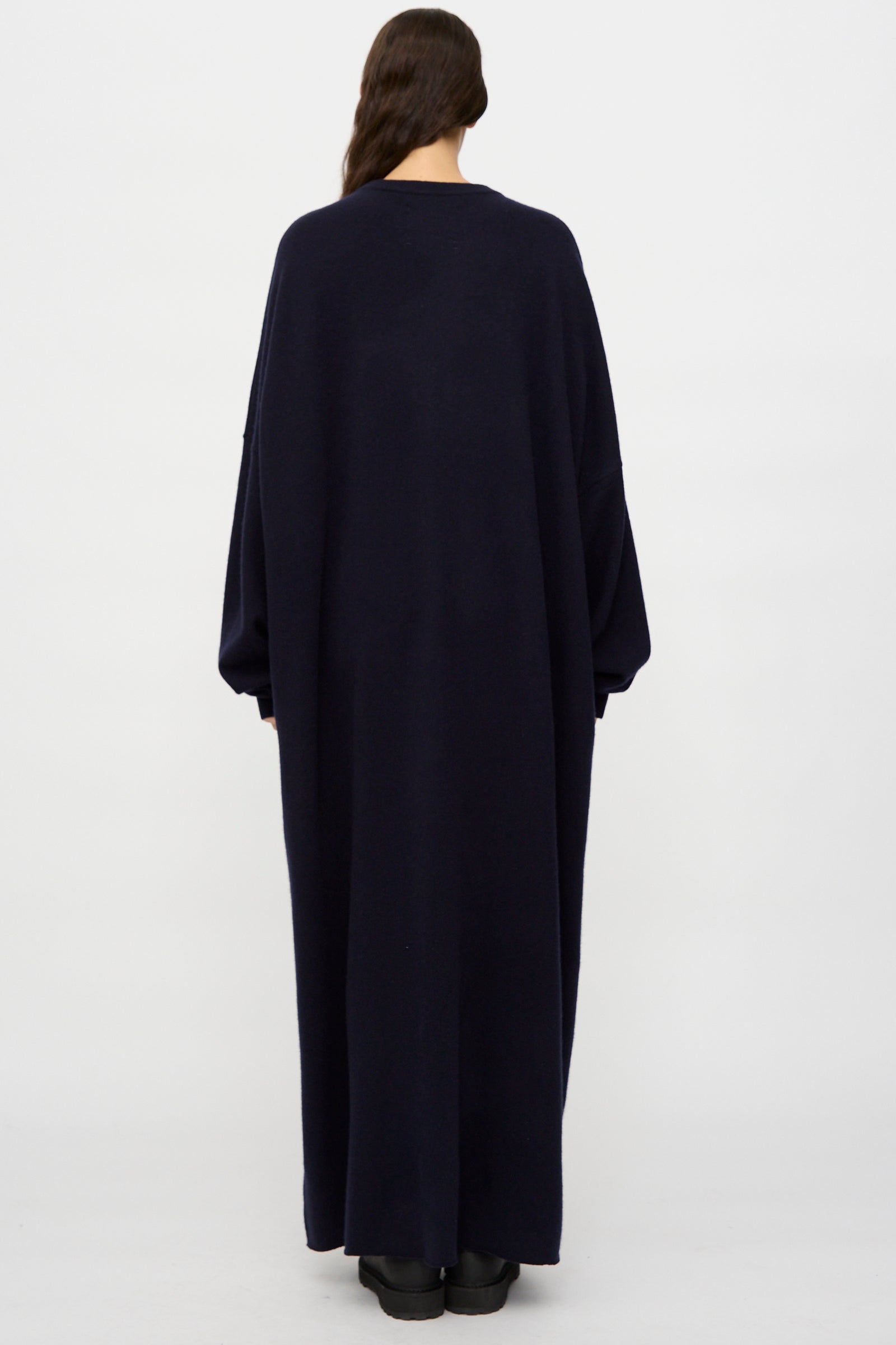 A person stands facing away, wearing the Extreme Cashmere No. 289 May Dress in navy—a loose-fit, long sleeve maxi dress—set against a plain white background.