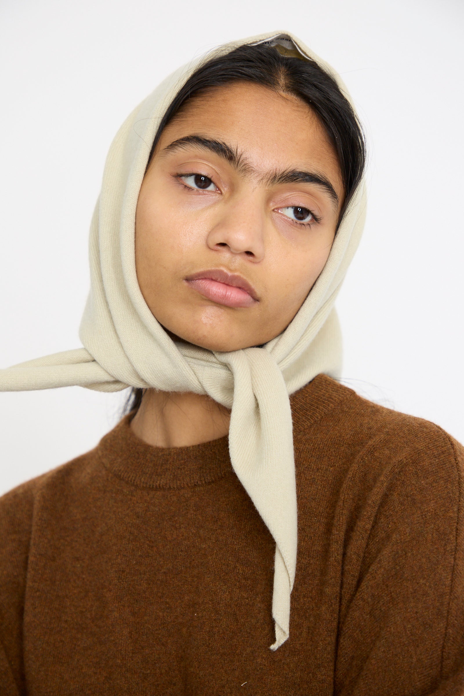 A model wears The No. 35 Bandana in Desert from Extreme Cashmere , a beige headscarf, and brown sweater, gazing sideways neutrally. 