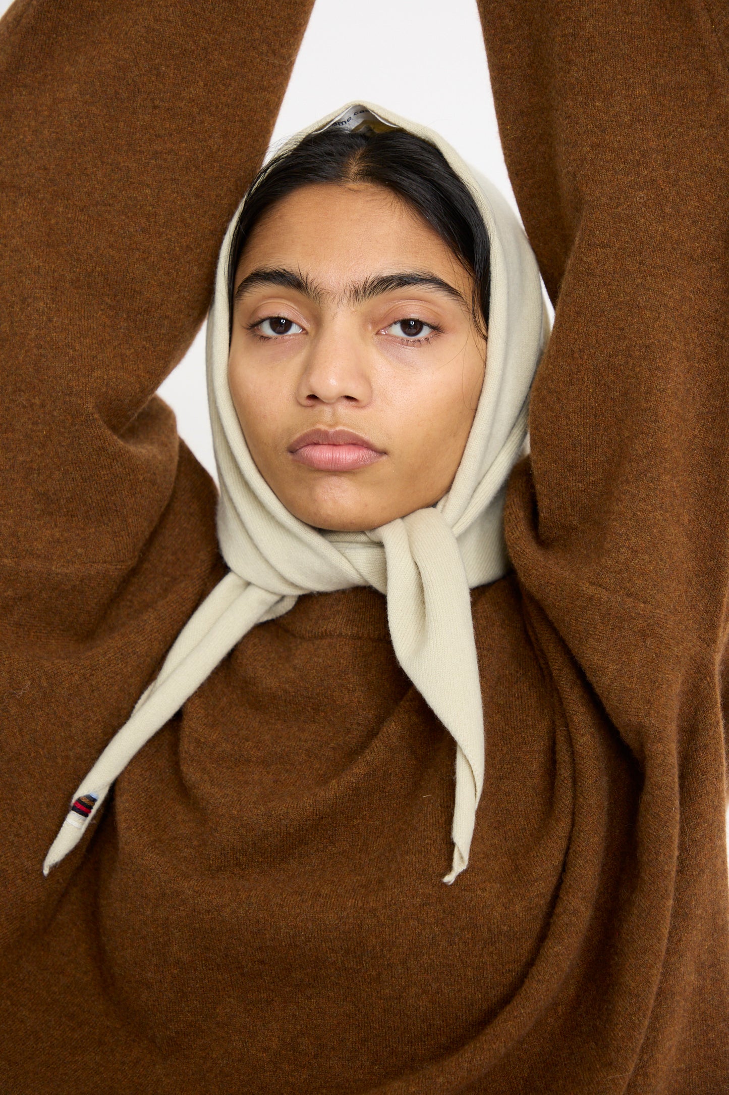 A model in a brown sweater and Extreme Cashmere's No. 35 Bandana in Desert, raises their arms overhead and looks forward. 