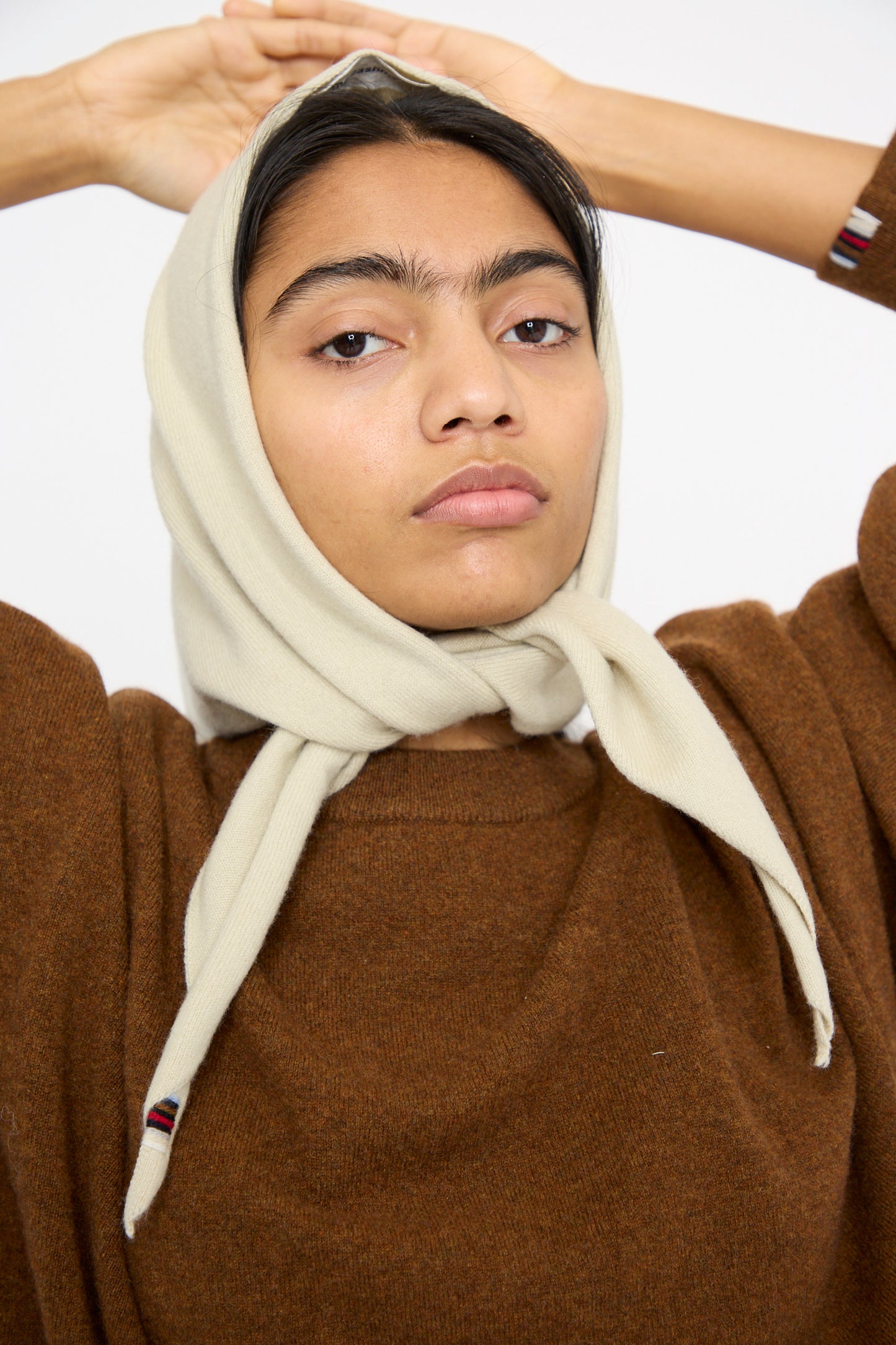 A model dressed in a soft brown sweater and Extreme Cashmere's No. 35 Bandana in Desert, gazes directly at the camera with arms elegantly raised behind their head.  