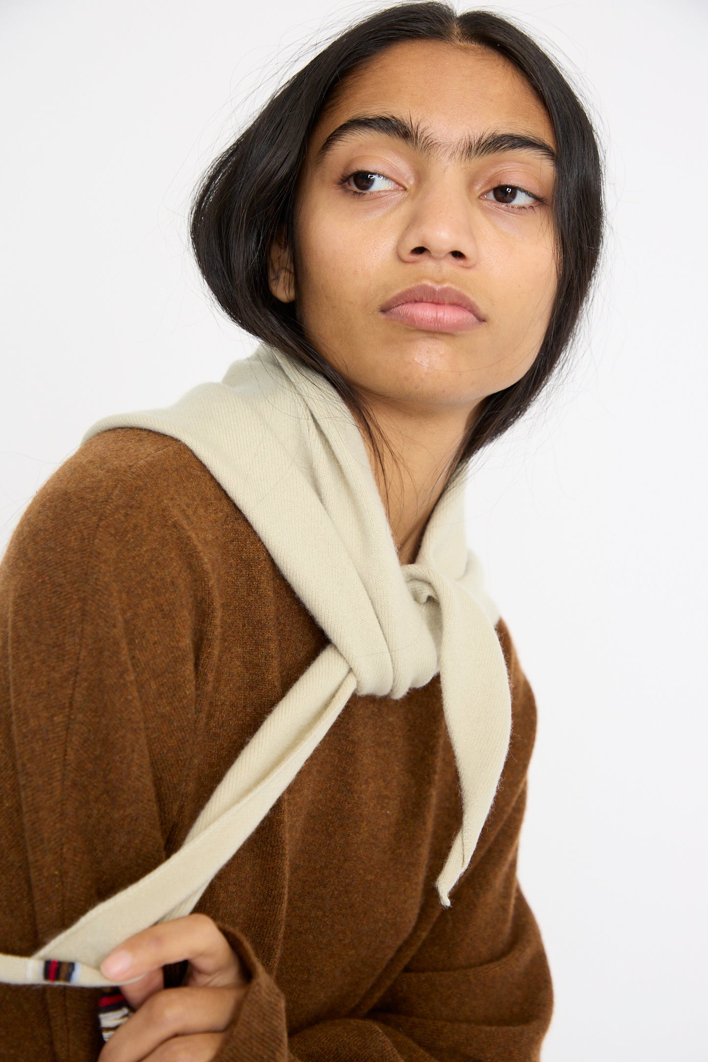 A person with long dark hair, wearing a brown sweater and Extreme Cashmere's No. 35 Bandana in Desert, looks to the side against a plain background.