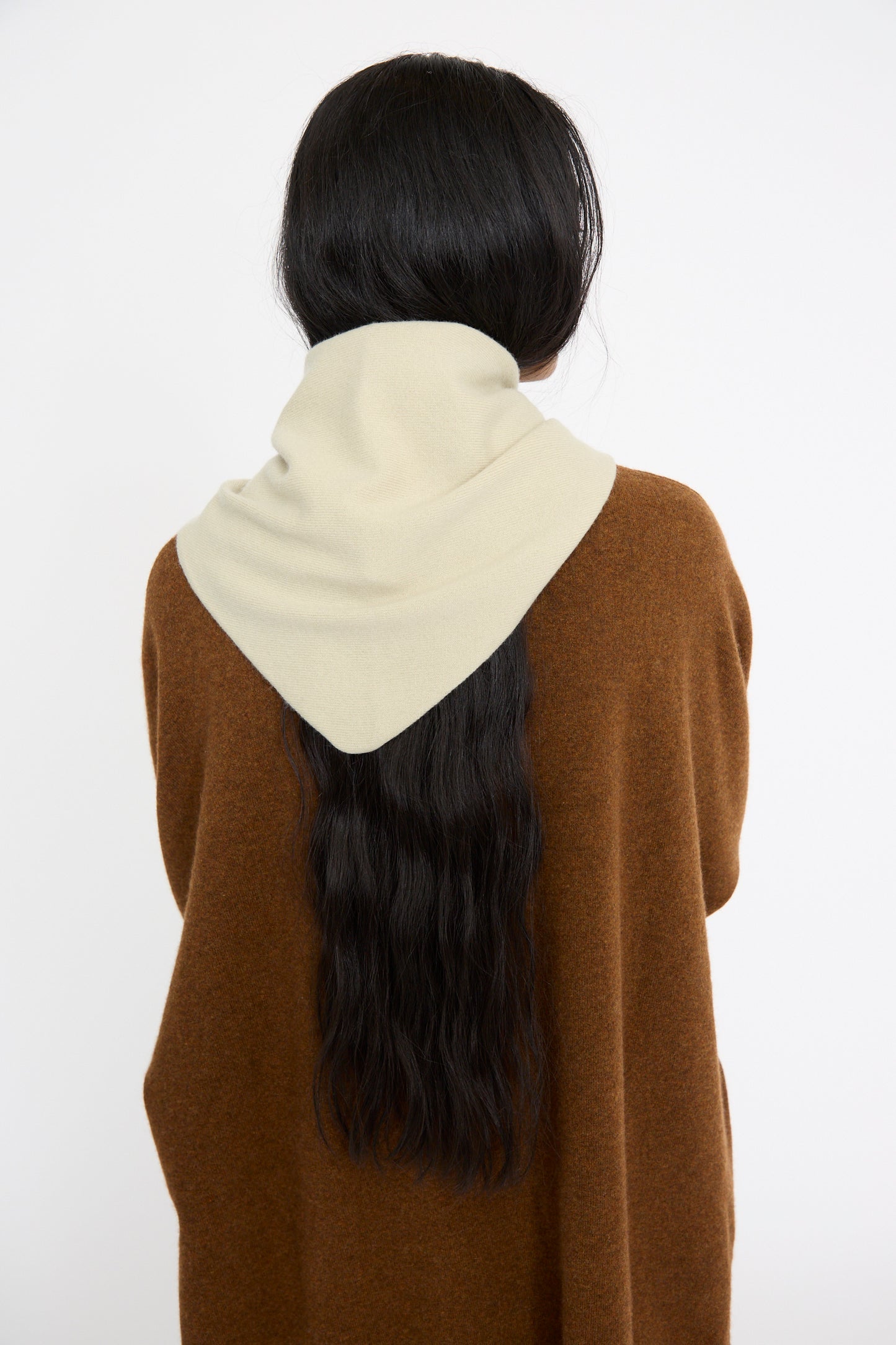 A person with long dark hair, wearing a brown sweater and Extreme Cashmere No. 35 Bandana in Desert is seen from behind against a plain background. 