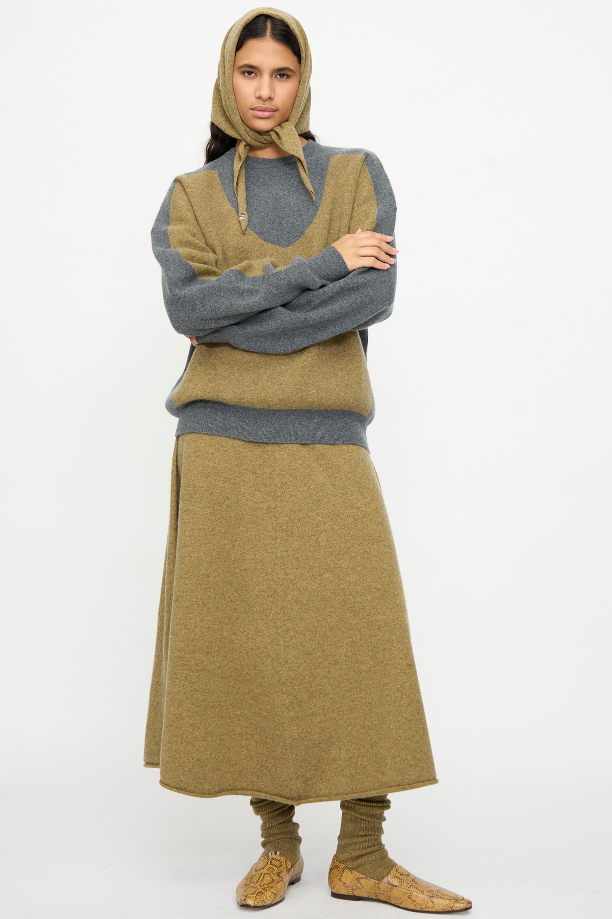 A person with arms folded across their chest stands against a white background wearing an Extreme Cashmere No. 35 Bandana in Hunter styled as a headscarf, paired with a two-tone gray and tan sweater, matching skirt, socks, and tan slip-on shoes.