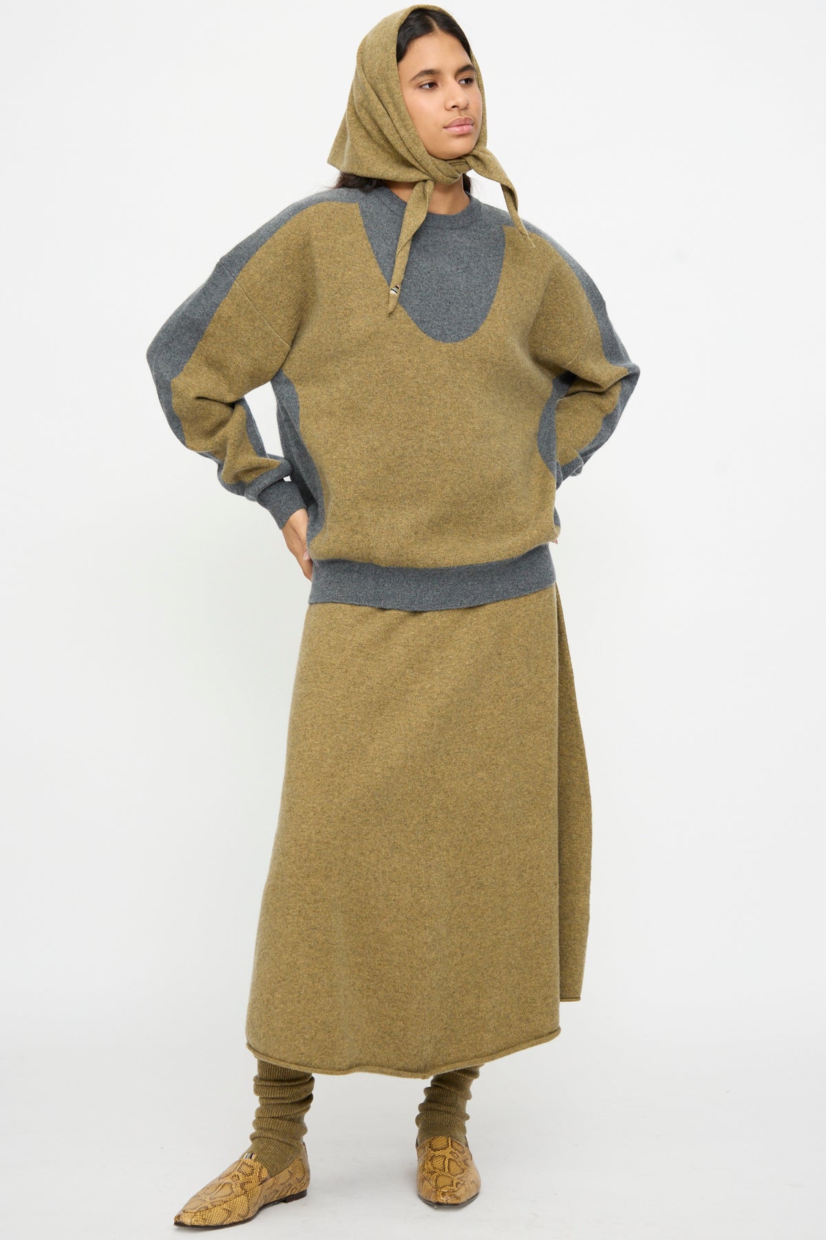A person wearing Extreme Cashmere’s No. 35 Bandana in Hunter stands against a plain white background, styled with a tan and gray hooded sweater, matching long skirt, coordinating leg warmers, and tan shoes.