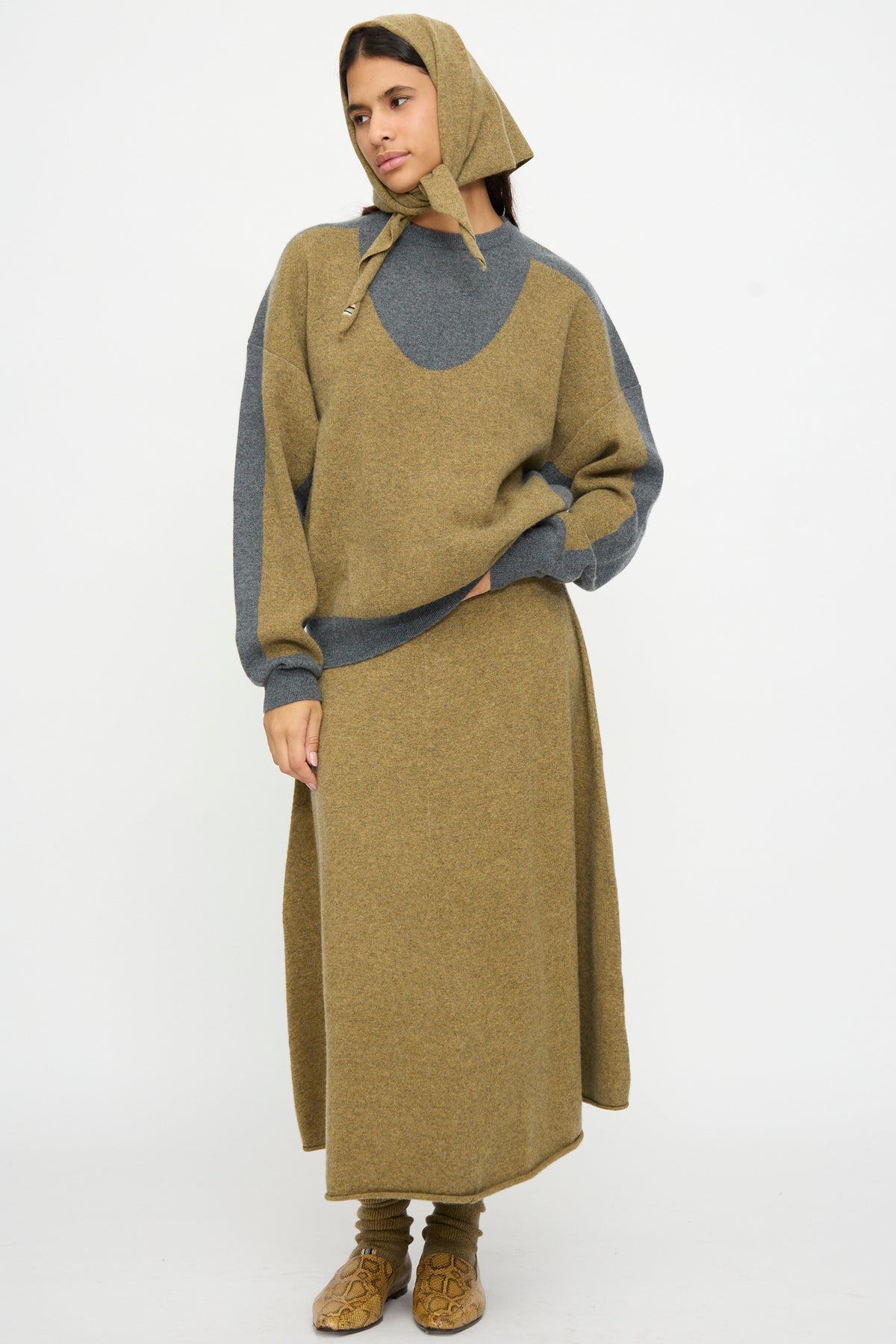 A person stands against a plain white background wearing an olive and gray knit hood, sweater, long skirt, and socks from Extreme Cashmere, accessorized with the No. 35 Bandana in Hunter.