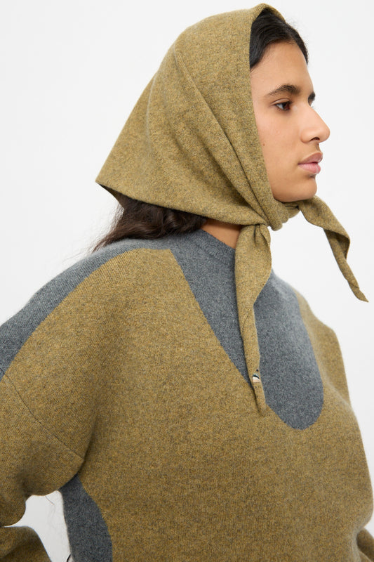 A person in profile wears the No. 35 Bandana in Hunter by Extreme Cashmere, tied under the chin, paired with a coordinating brown and gray sweater, standing against a plain white background.
