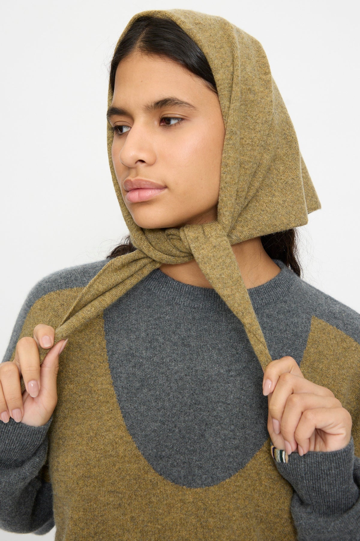 A person wears the Extreme Cashmere No. 35 Bandana in Hunter, styled as a headscarf, along with a gray and olive sweater, gazing to the side against a plain background.