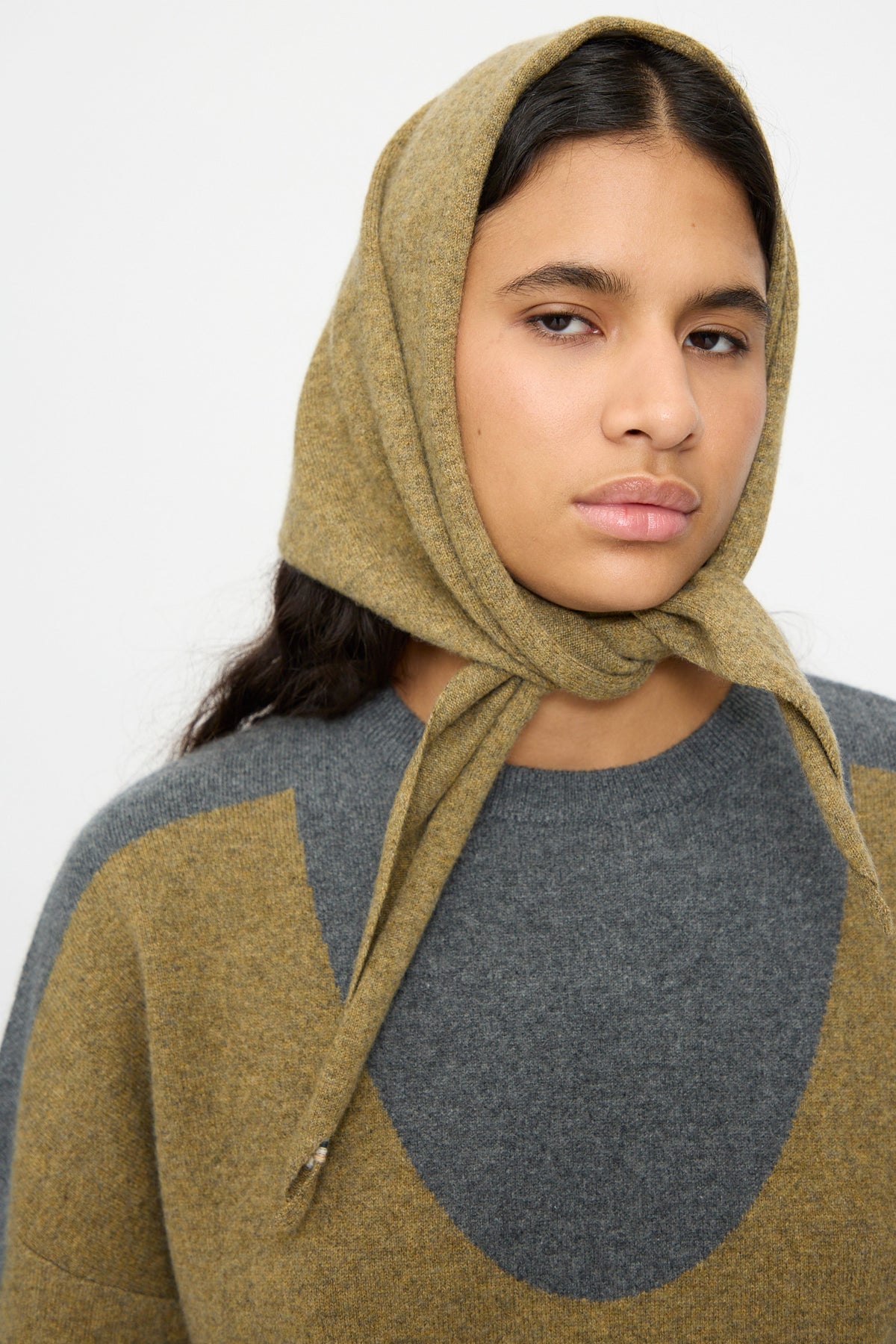 A person wearing a gray and olive green sweater and styling the Extreme Cashmere No. 35 Bandana in Hunter as a headscarf, looking at the camera against a plain background.