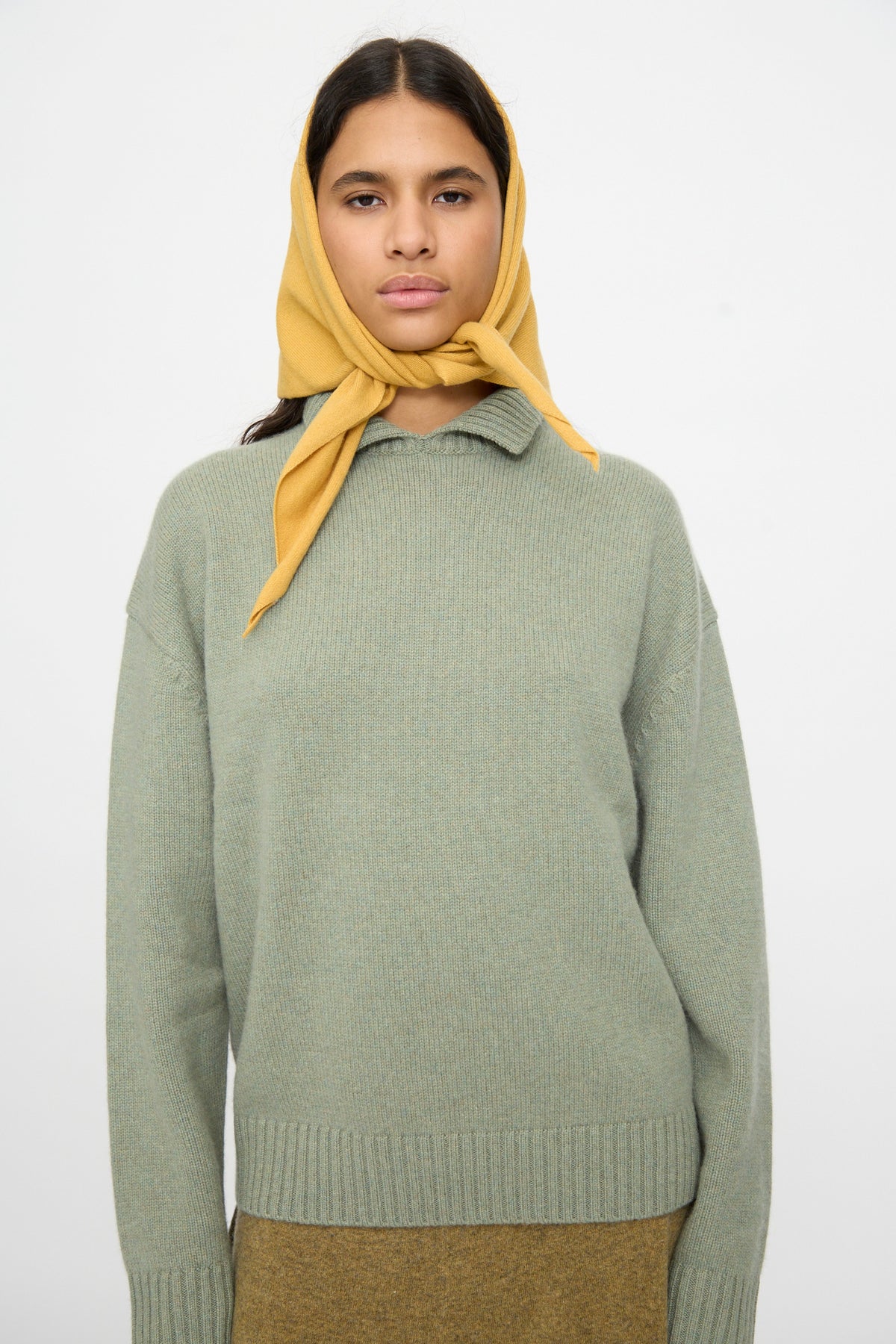 A person wearing a green knit sweater and the Extreme Cashmere No. 35 Bandana in Wheat stands against a plain white background, looking forward.